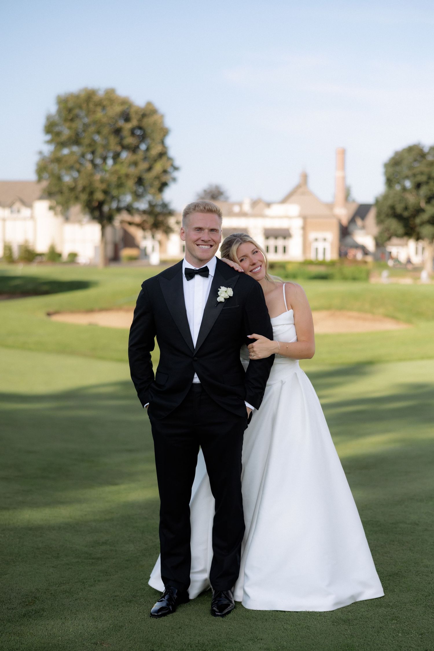 Smiling bride and groom pose together on golf course, bride in white gown, groom in black tuxedo.