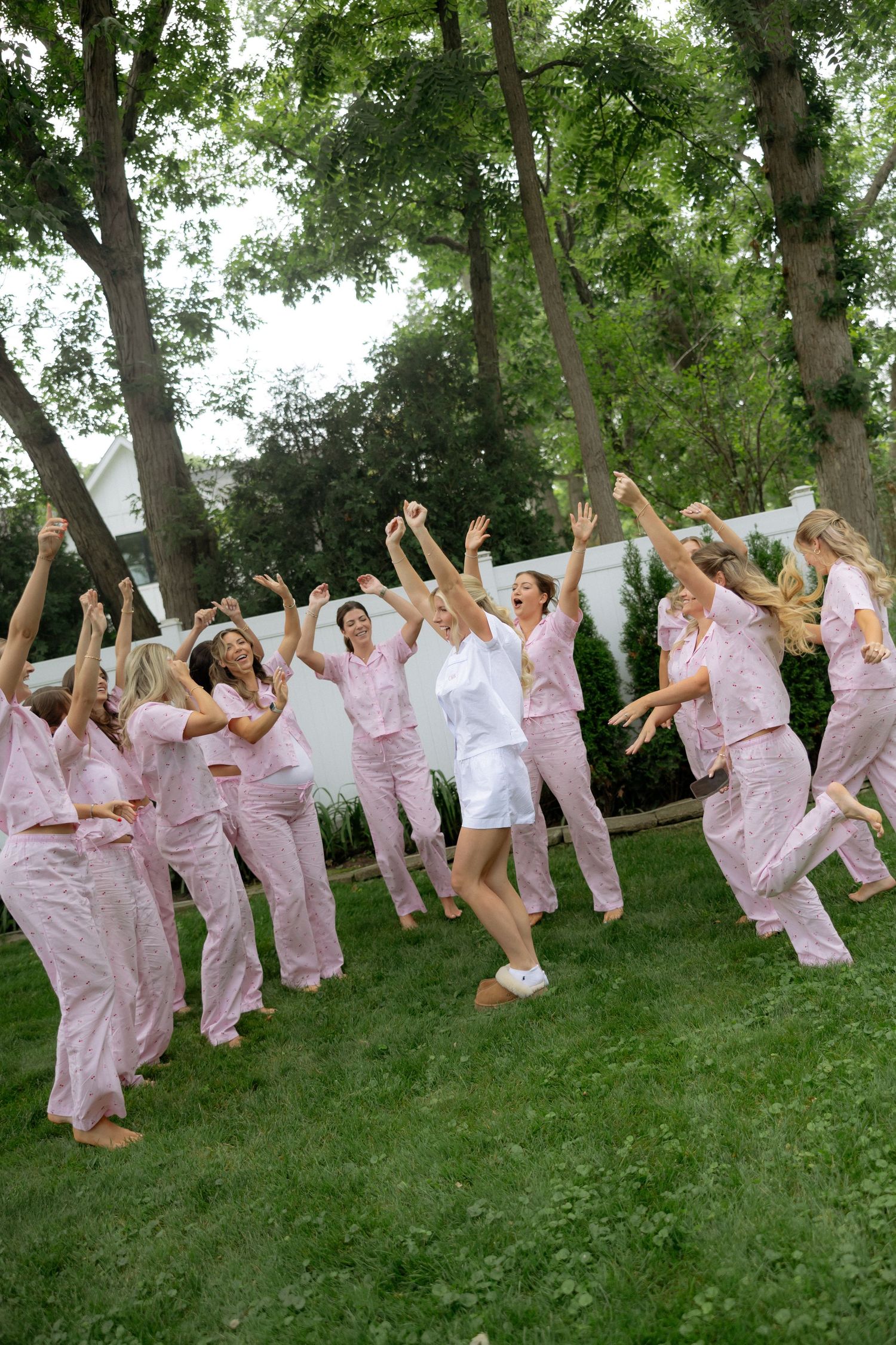A bride in white and bridesmaids in pink pajamas celebrate joyfully outdoors on green grass with arms raised.