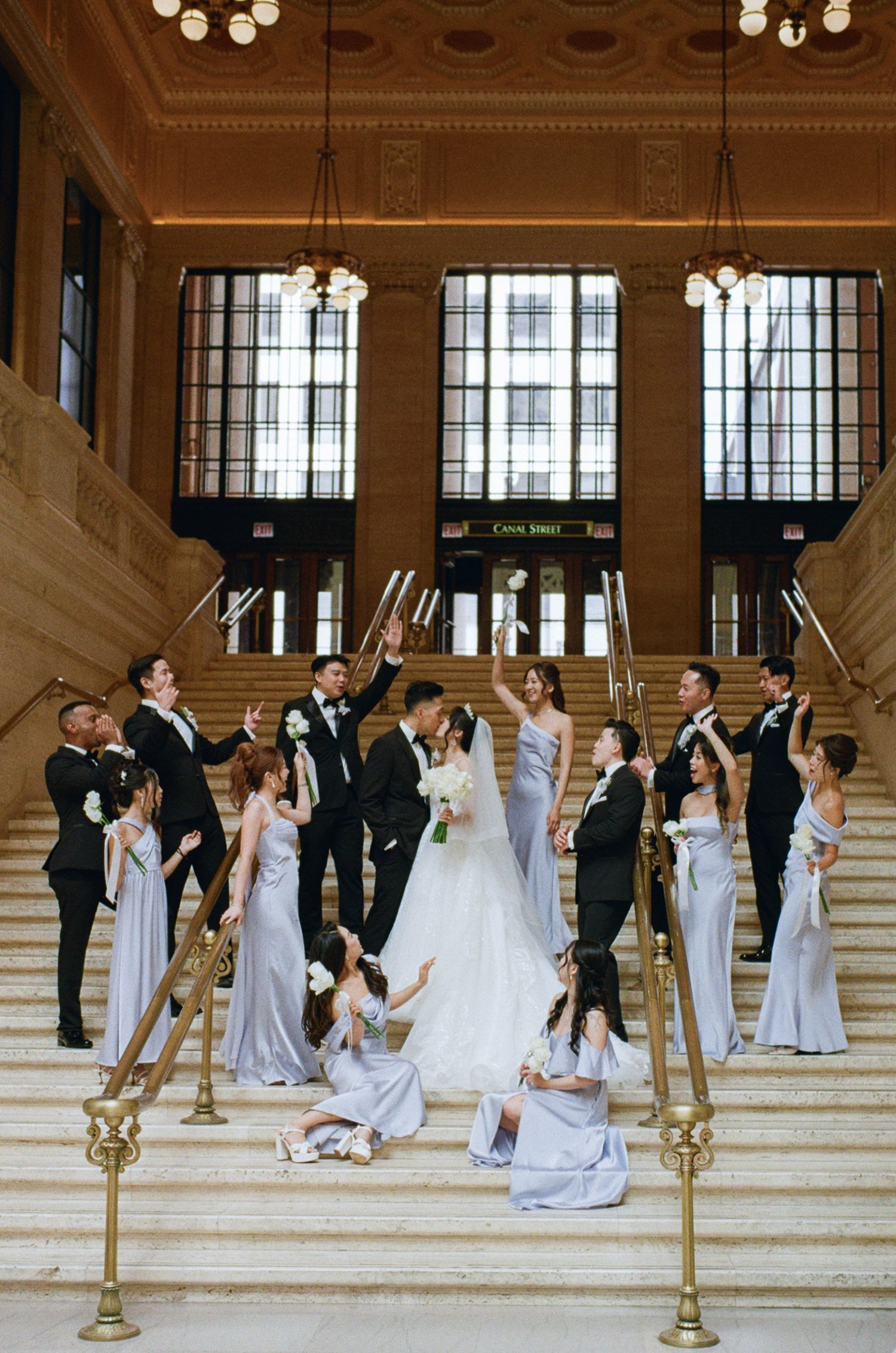 Wedding party celebrates on grand staircase, bride and groom kissing, bridesmaids in lavender gowns, groomsmen in black suits.