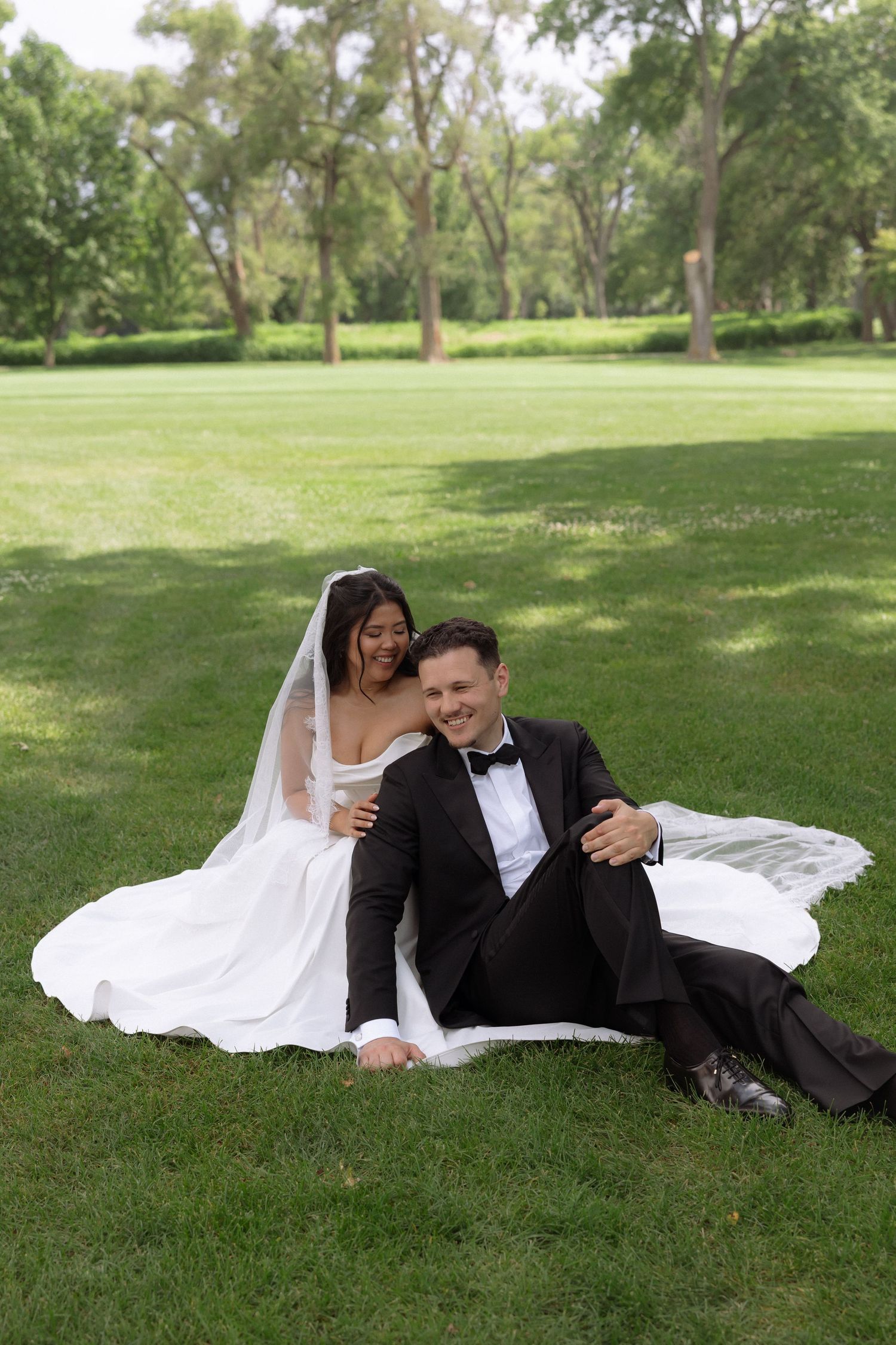 Happy bride and groom sitting together on green grass laughing in a scenic park setting.