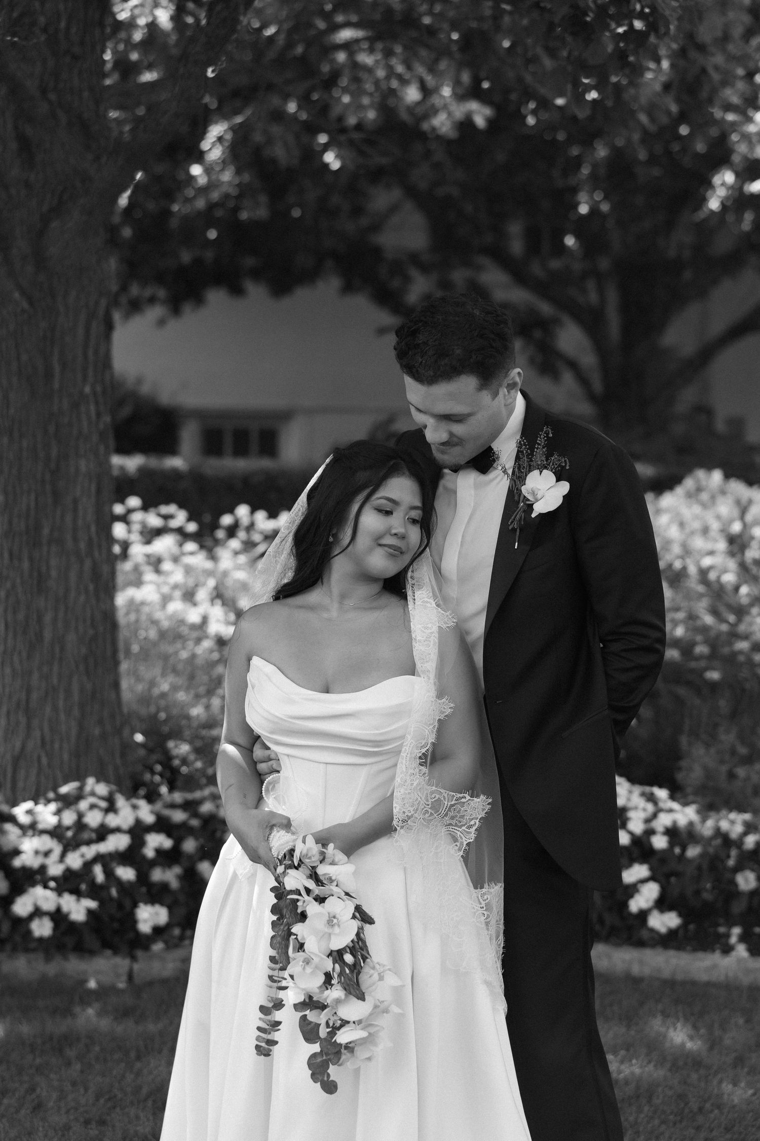 Bride and groom share tender embrace near flowering garden in elegant black and white portrait.