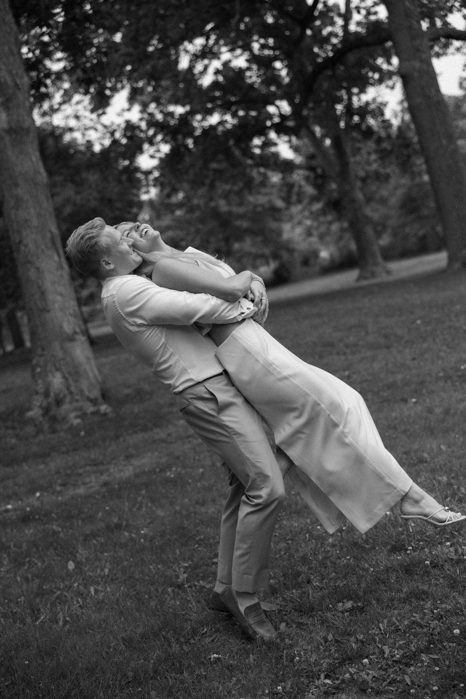 A joyful couple shares a playful lift in a tree-lined park in a classic black and white photo.