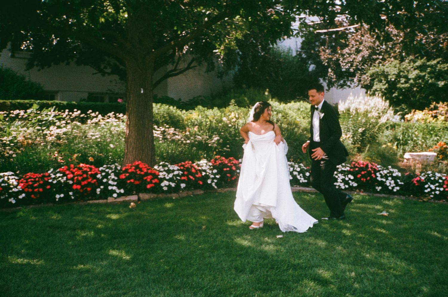 Bride and groom walking hand in hand on lush green lawn surrounded by colorful flower gardens on their wedding day.