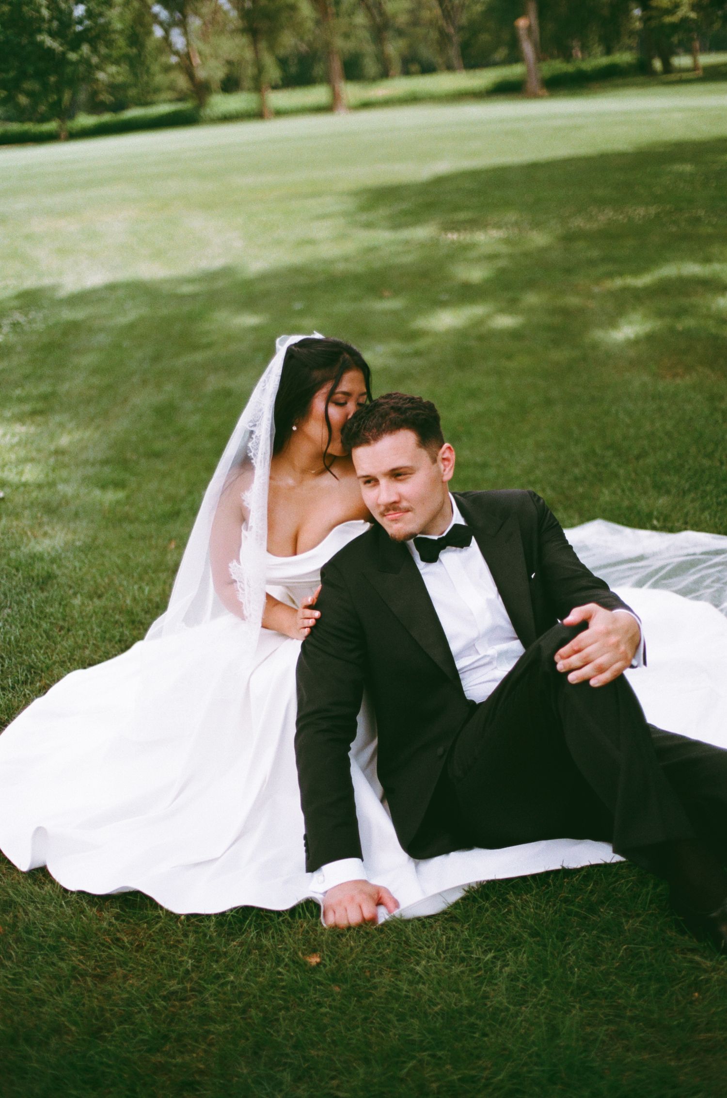 Bride and groom sitting on lush green grass, bride in white gown with veil, groom in black tuxedo with bow tie.