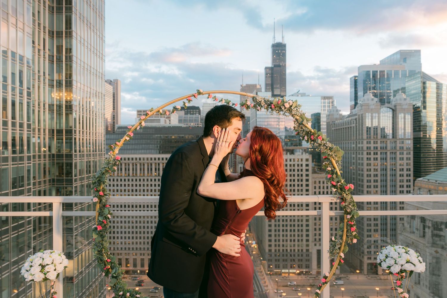 Couple shares a tender kiss after rooftop proposal with Chicago skyline and Willis Tower in background.