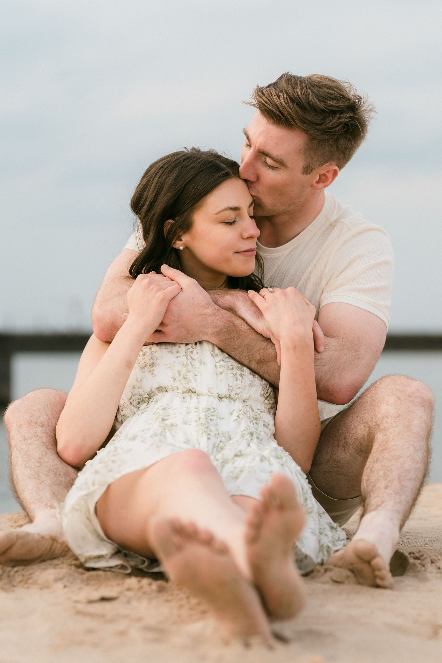 Couple sitting on sandy beach, man kissing woman on forehead while gently embracing her from behind.