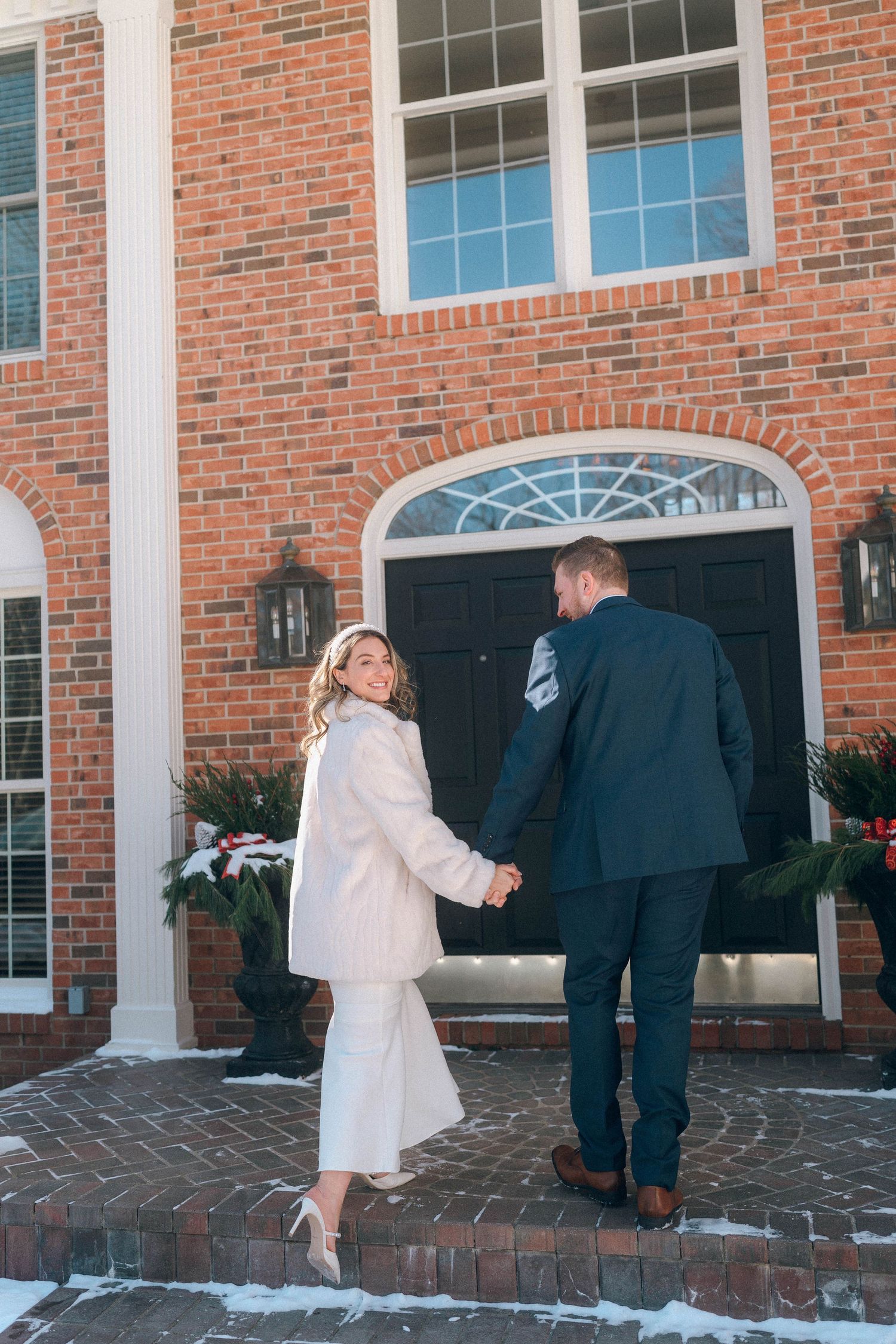Bride in white fur coat holds groom's hand walking toward brick mansion entrance in winter snow.