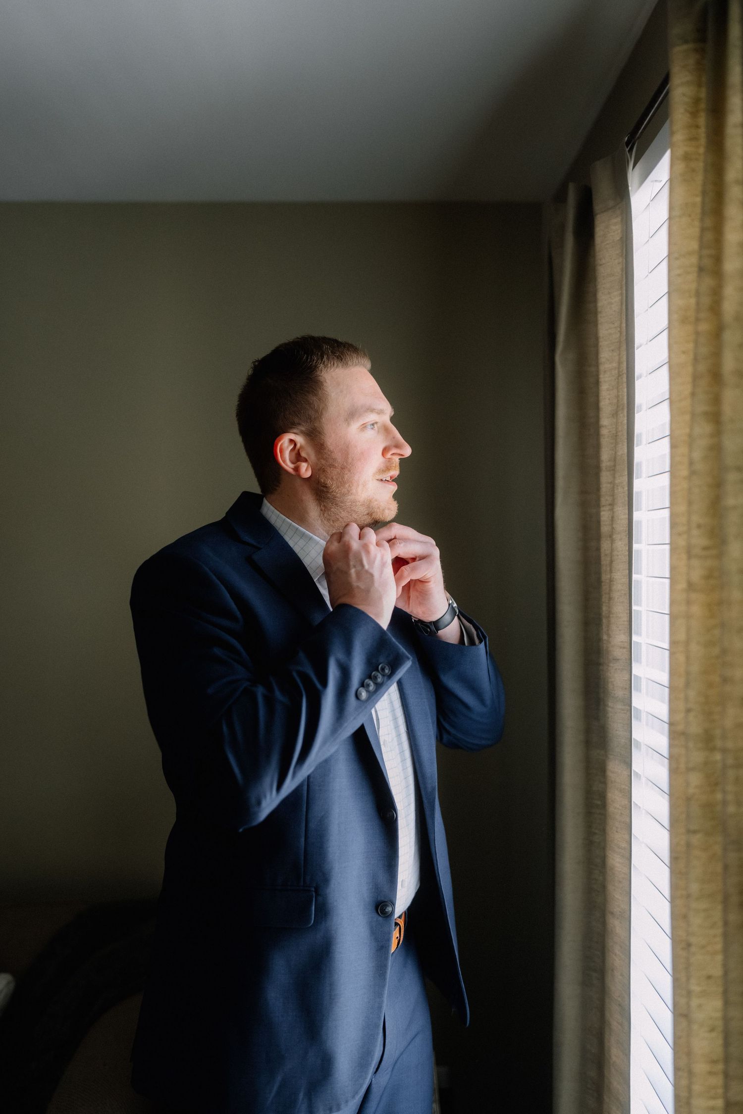 Handsome groom in navy suit adjusting collar by window before wedding ceremony.
