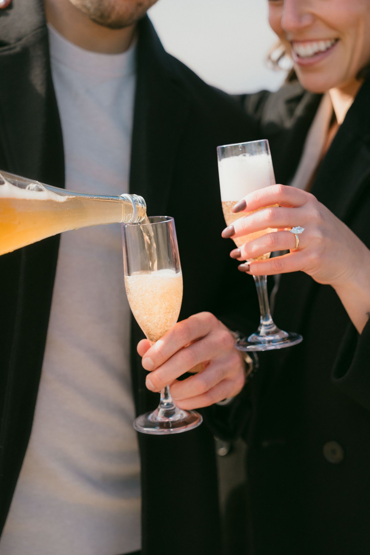 Close-up of champagne being poured into flutes held by an engaged couple with a diamond ring.