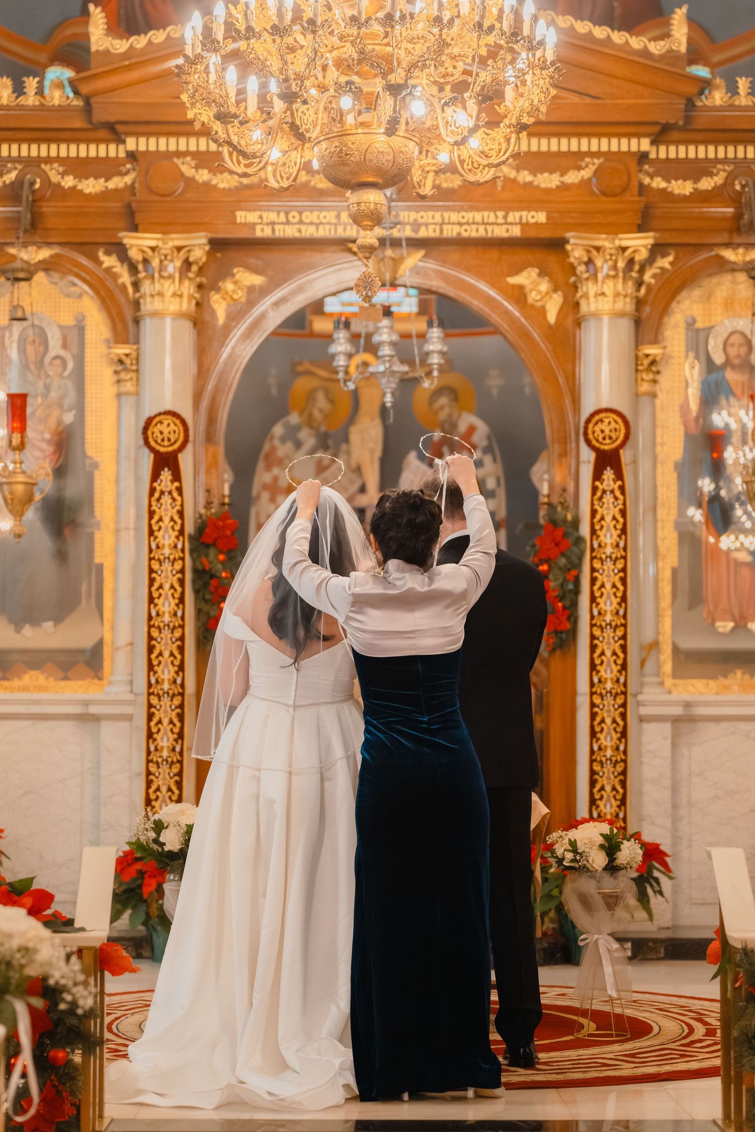 Orthodox priest crowns bride and groom during traditional Greek wedding ceremony at ornate golden iconostasis altar.