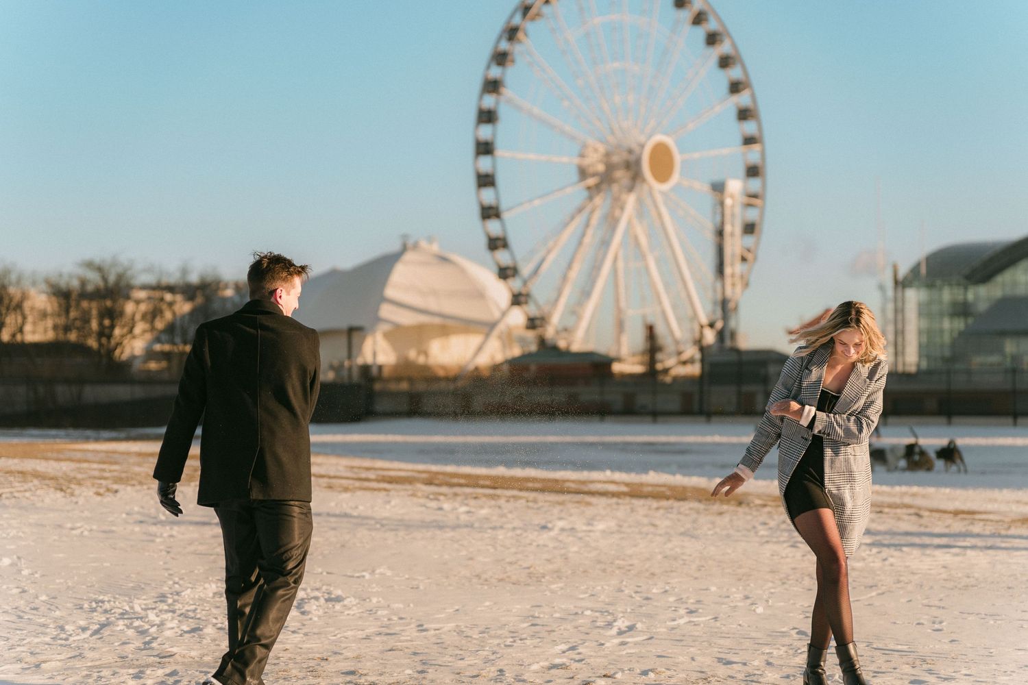 A couple walks apart playfully on a snowy open field with a Ferris wheel in the background.