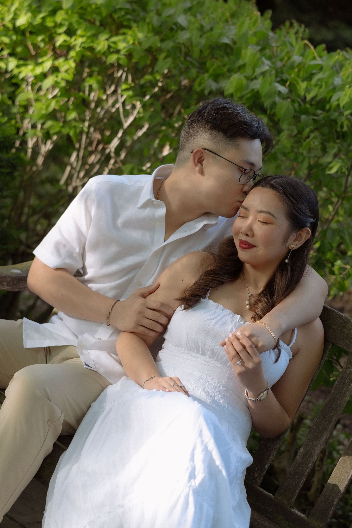 A romantic Asian couple in white outfits share a tender kiss outdoors, surrounded by lush green foliage.