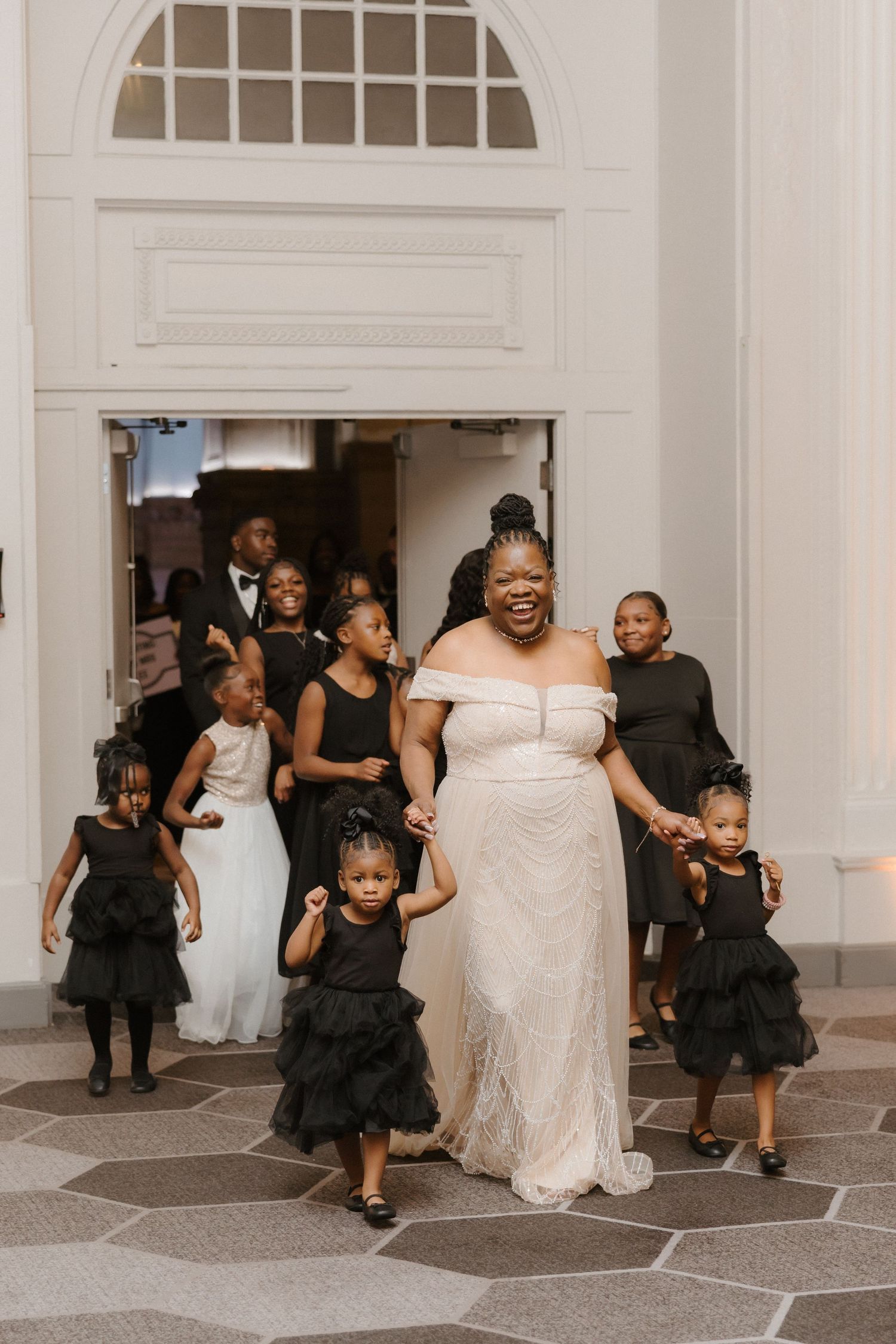 Bride in white off-shoulder gown exits church with flower girls in black dresses, smiling joyfully.