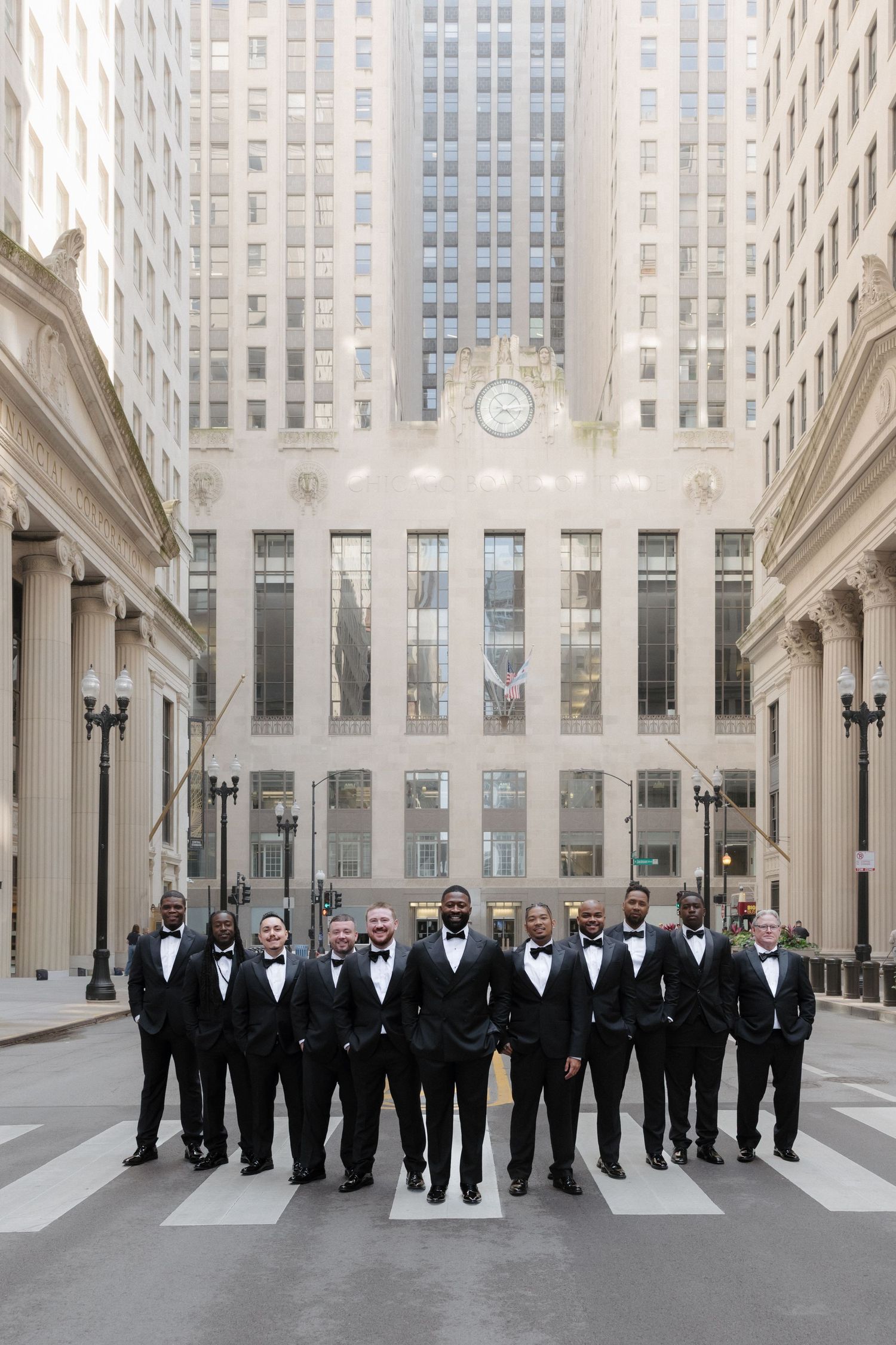 Groomsmen in black tuxedos and bow ties posing on a crosswalk in an urban cityscape with classic architecture.