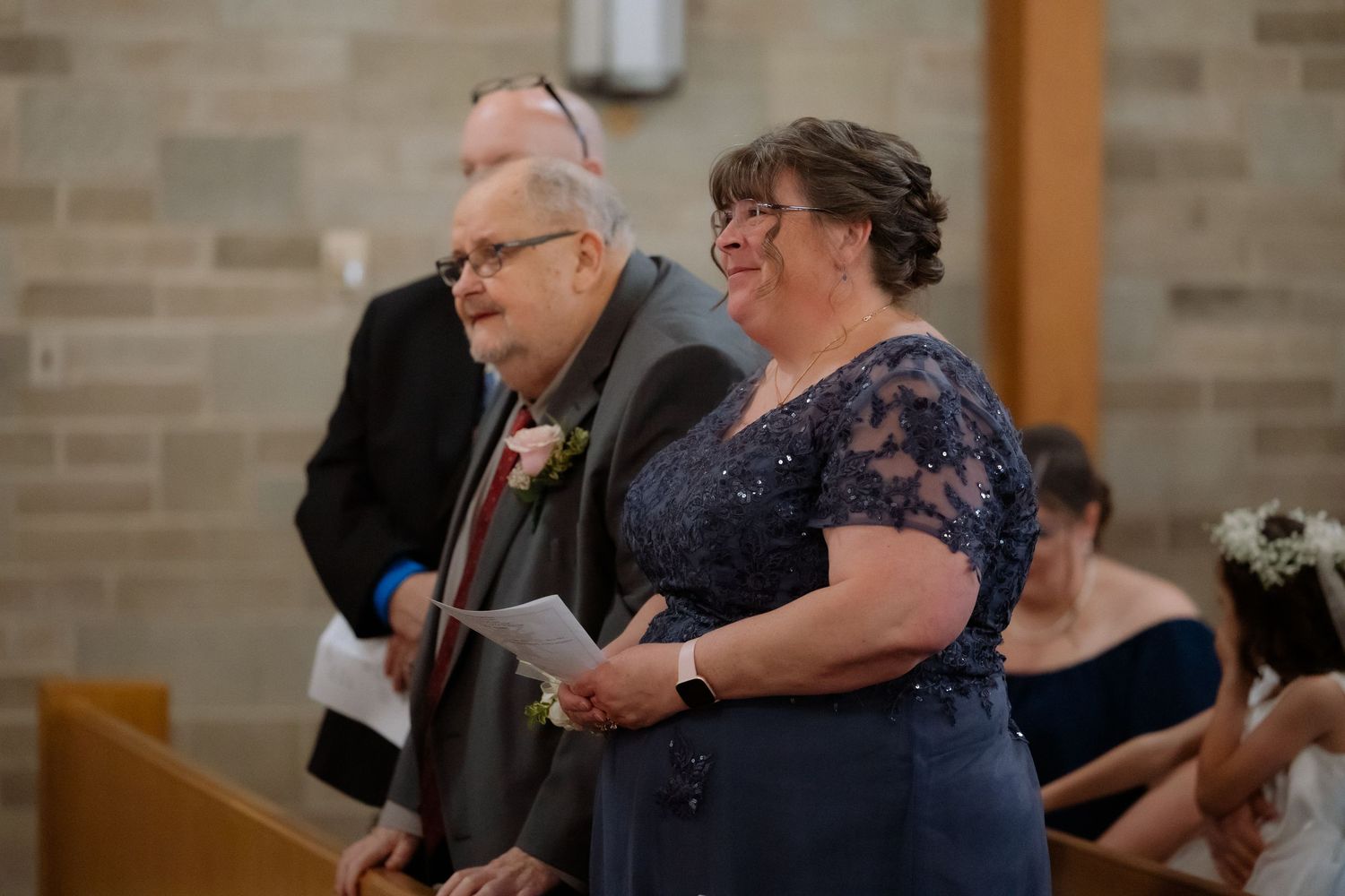 Elderly couple standing together in a church during a wedding ceremony, woman in blue lace dress holding a paper.