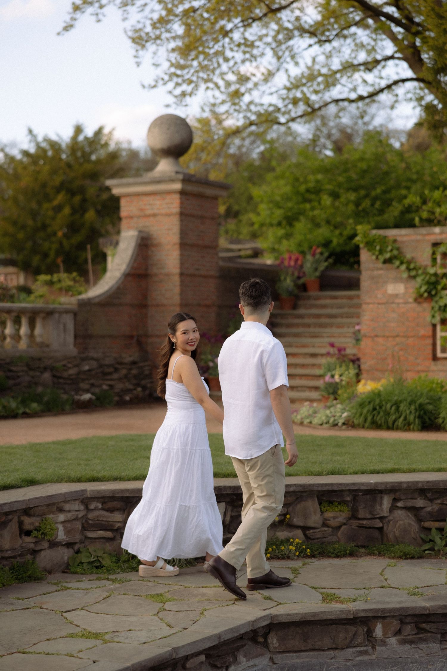 A couple in white outfits walks hand-in-hand through a lush garden with brick architecture and stone pathways.