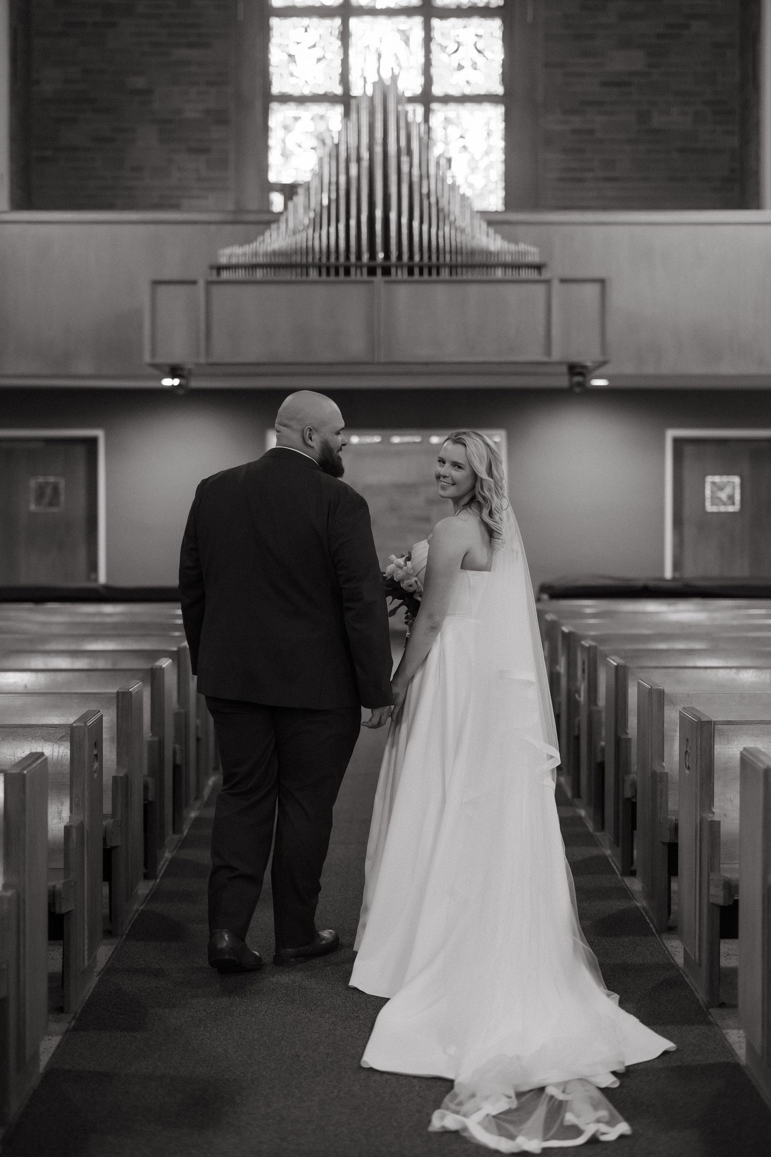 Black and white photo of a bride and groom holding hands in a church aisle with organ pipes in the background.