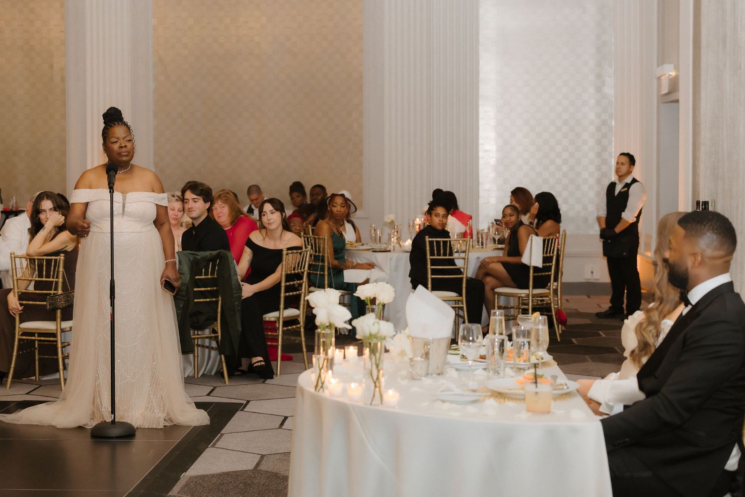 Woman giving a speech at an elegant wedding reception in a grand ballroom with candles and floral centerpieces.