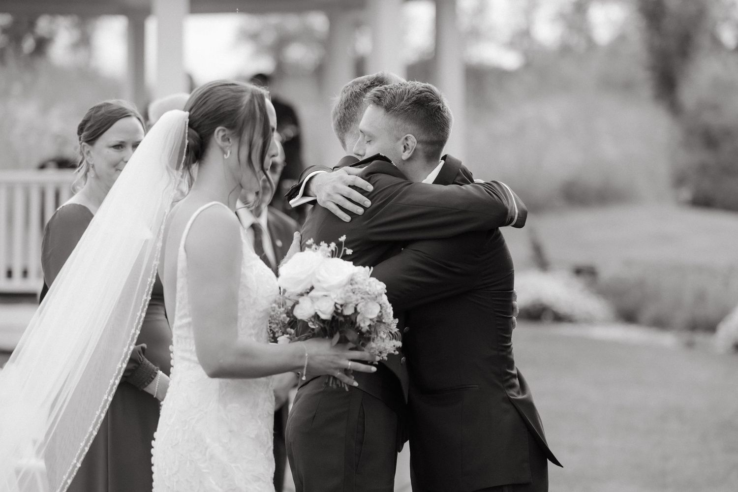 Black and white photo of bride embracing someone at outdoor wedding ceremony while holding white bouquet.