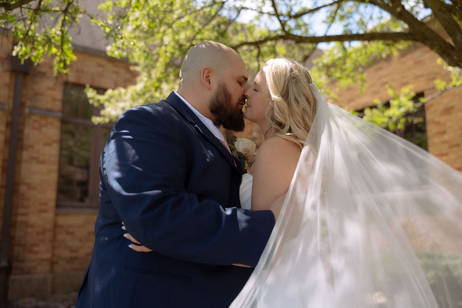 A bride and groom share a romantic kiss outdoors, her veil flowing in the breeze, with a brick building and trees behind.