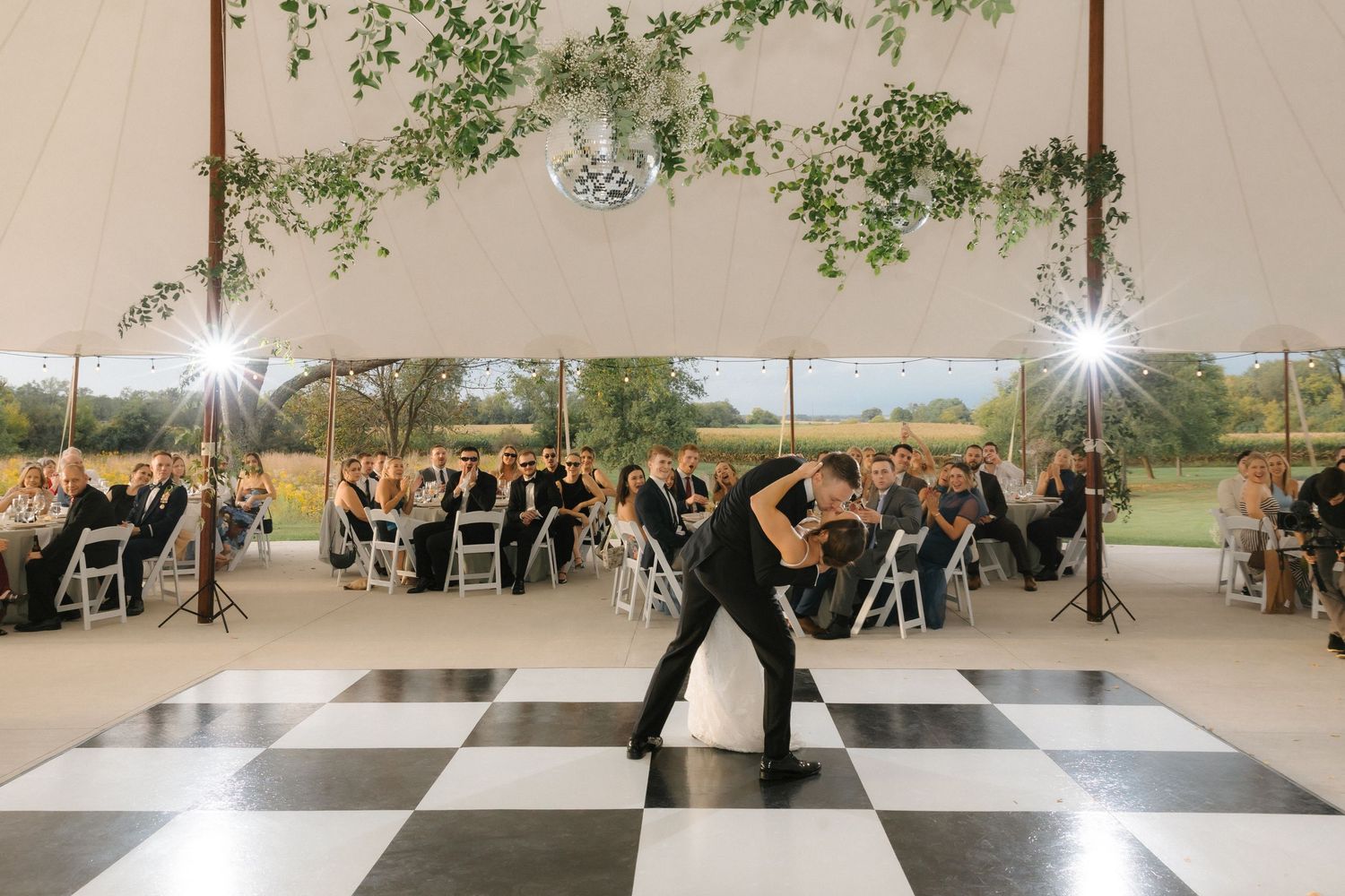 Groom dips bride dramatically on a black and white checkered dance floor while wedding guests watch in a tent.