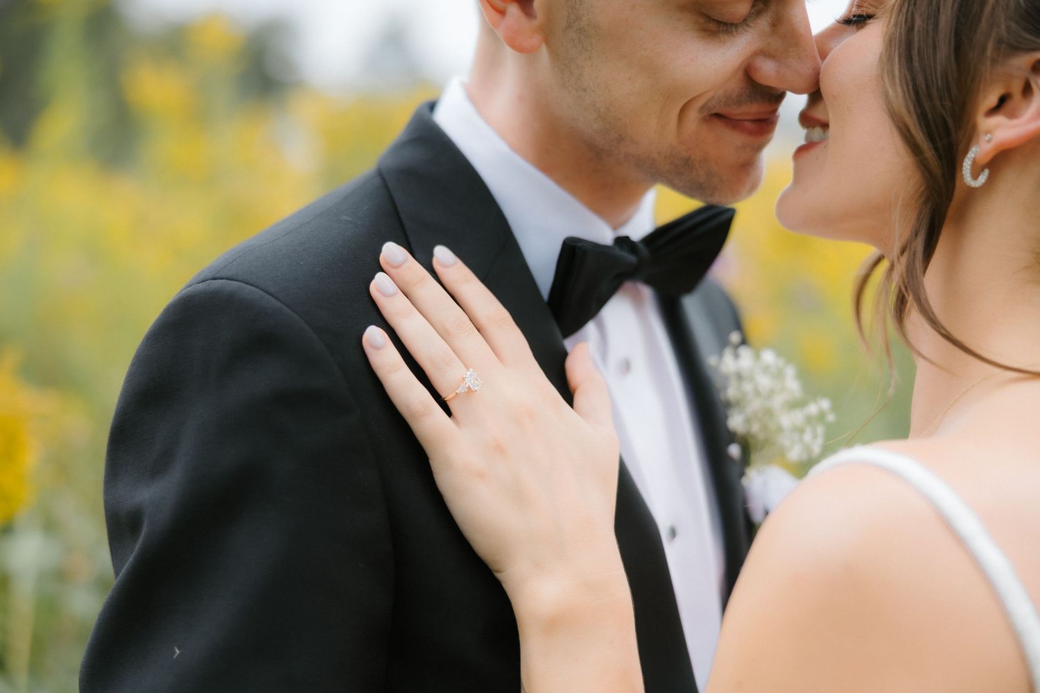 Bride and groom share a romantic almost-kiss, bride's hand resting on groom's chest showing wedding ring, yellow flowers behind.