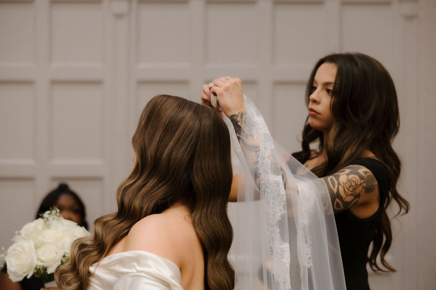 A bridesmaid with tattoos carefully adjusts a lace wedding veil on a bride holding white flowers.