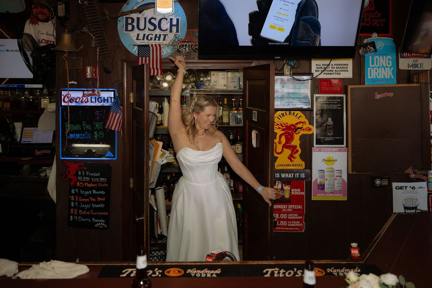 Bride in white dress celebrating behind a bar with Busch Light and Tito's signs in a festive bar setting.