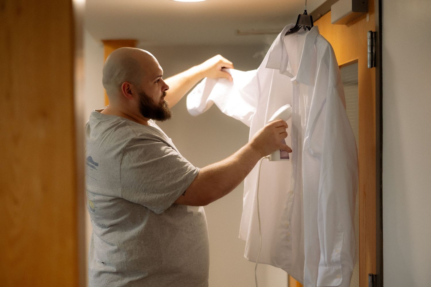 Bald bearded man selecting white dress shirts from a closet rack in a modern bedroom.