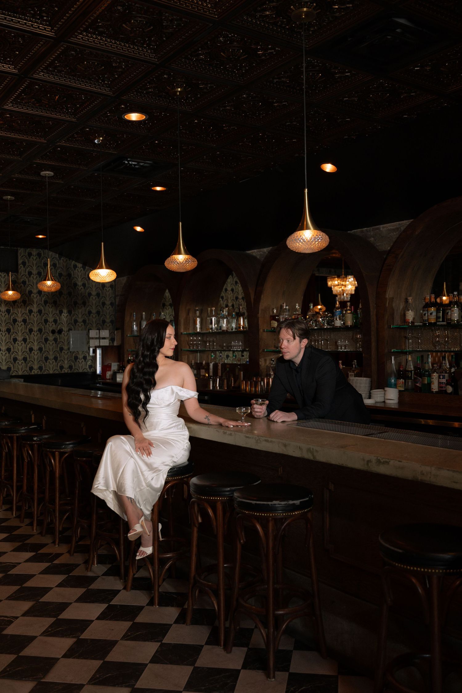 Couple in wedding attire sit together at a moody upscale bar with arched wood details and pendant lights.