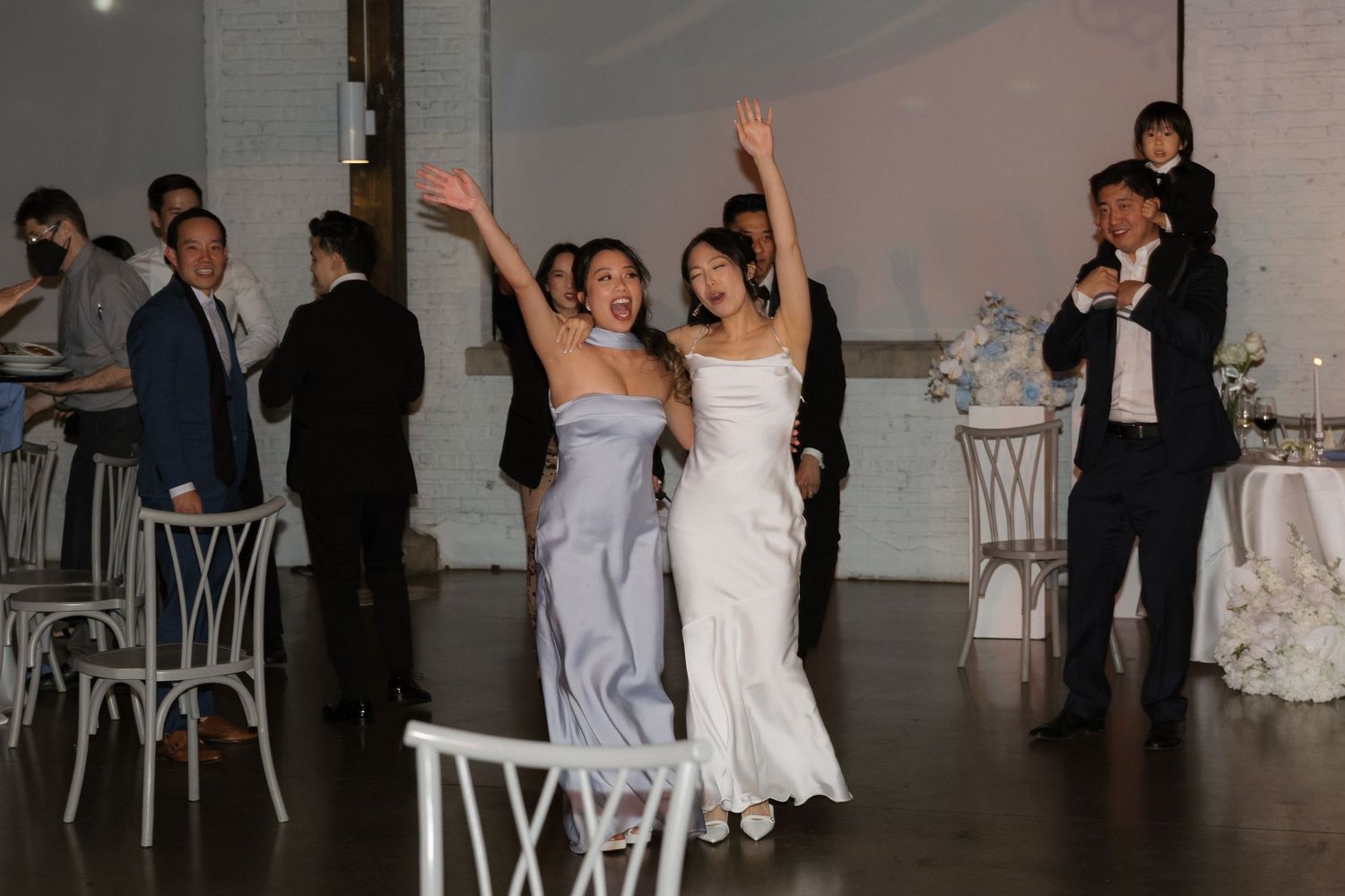 Bride in white gown and bridesmaid in lavender dress waving as they enter wedding reception.