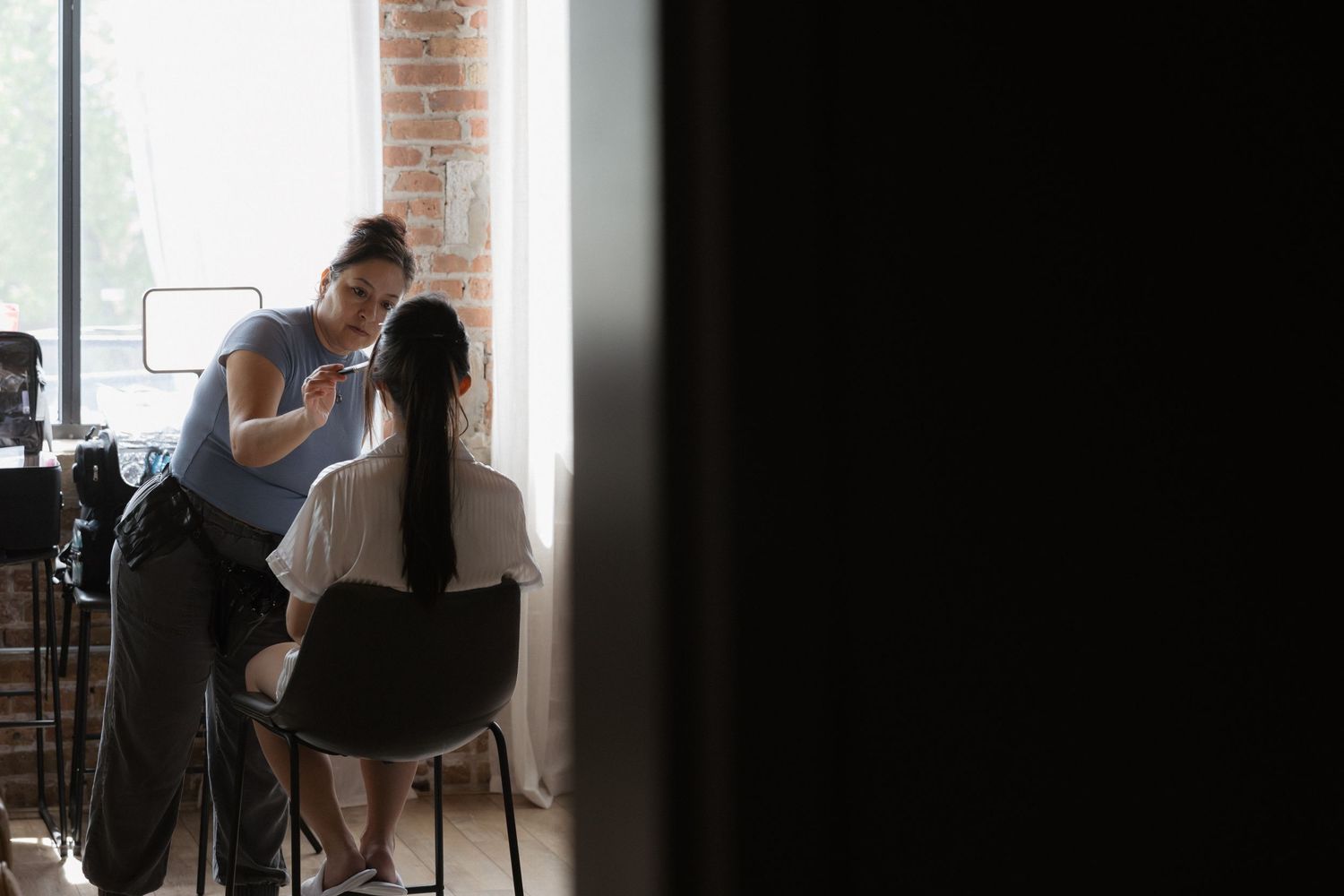A makeup artist applies makeup to a client seated in a chair near a bright window in a modern studio.