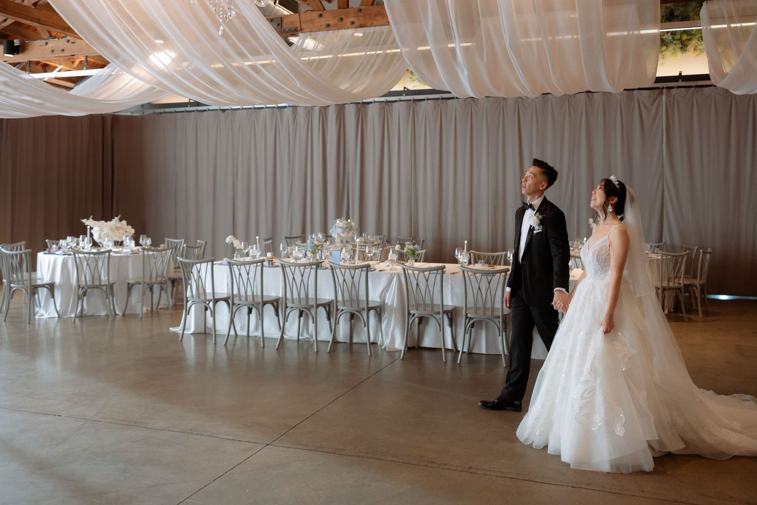 Bride and groom in elegant attire admiring beautifully decorated wedding reception venue with draped ceilings.