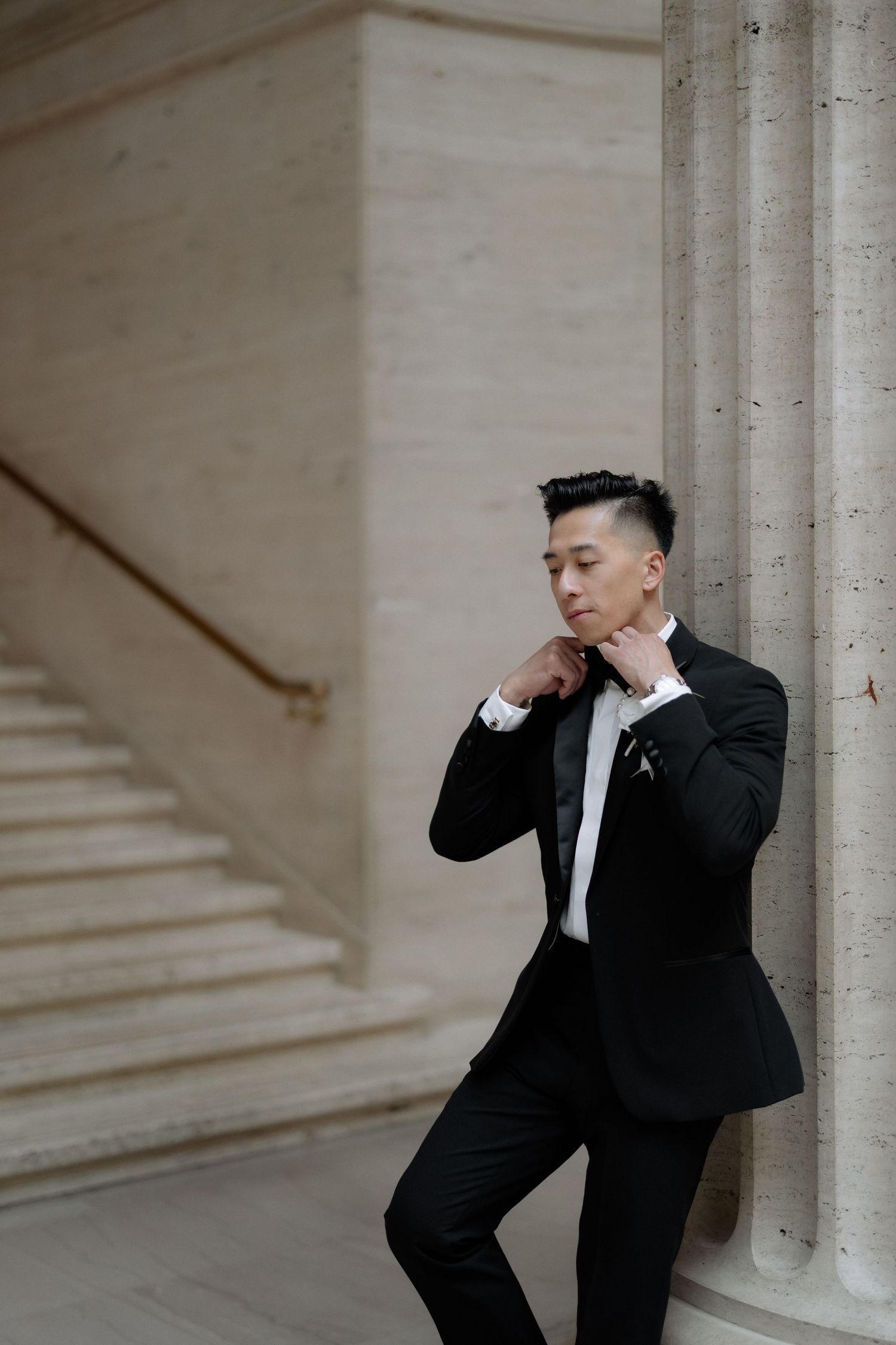 Elegant groom in black tuxedo adjusting his collar while leaning against a marble column.