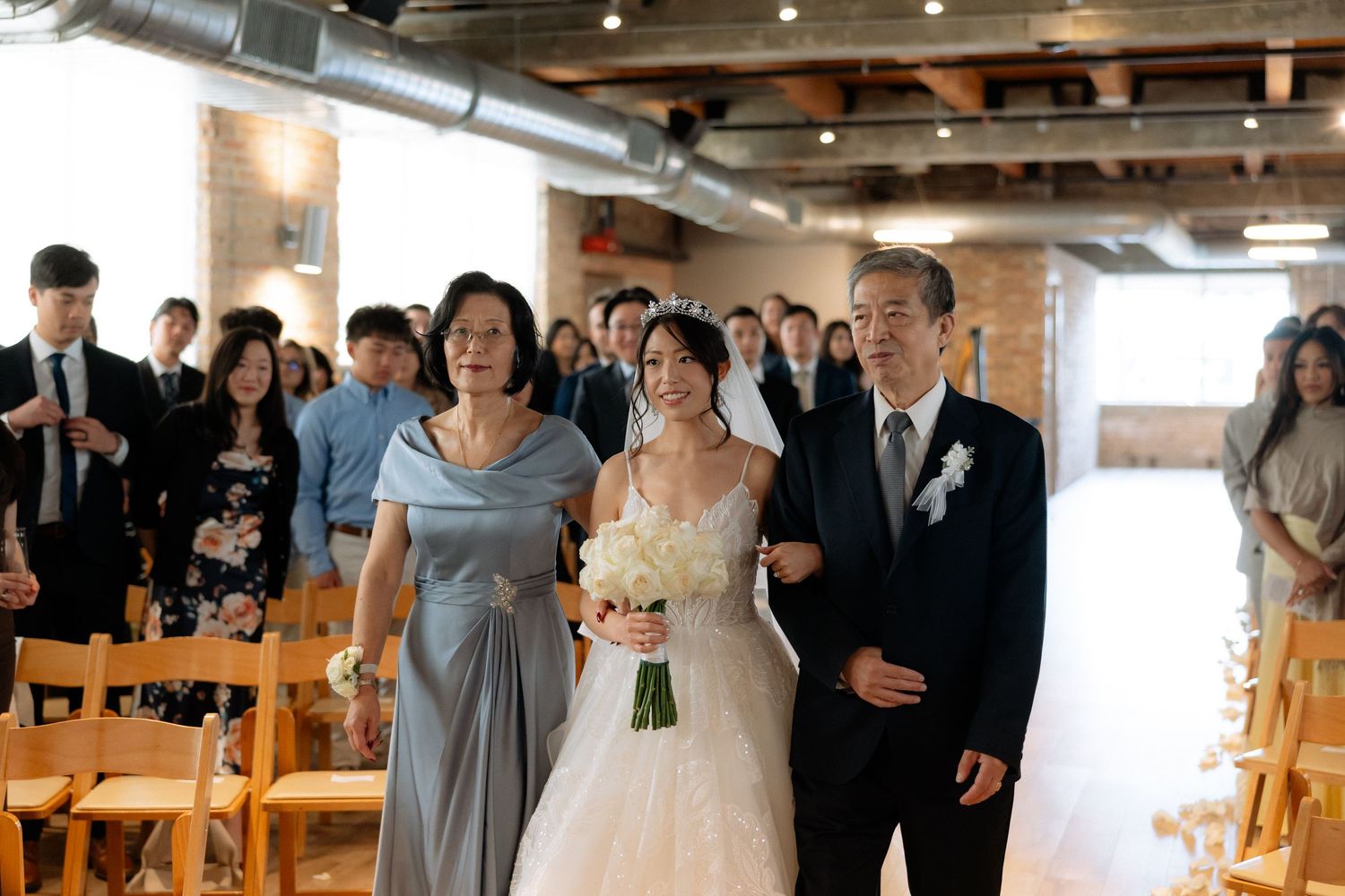 Bride walks down the aisle with parents during indoor wedding ceremony, holding white bouquet in elegant white gown.