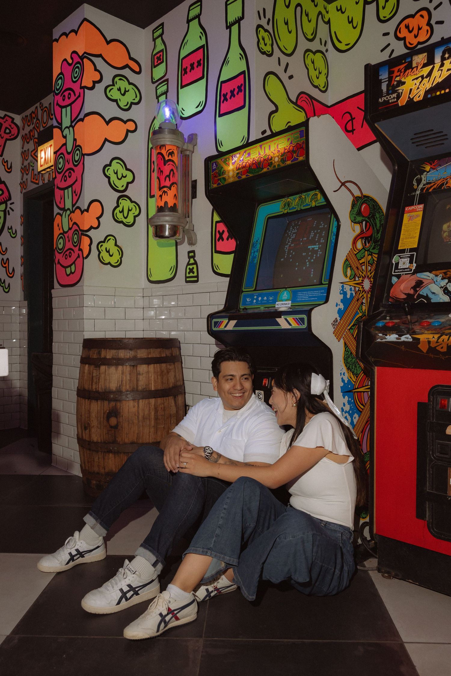 Couple sitting on floor by vintage arcade machines with colorful graffiti wall art in a retro game bar.