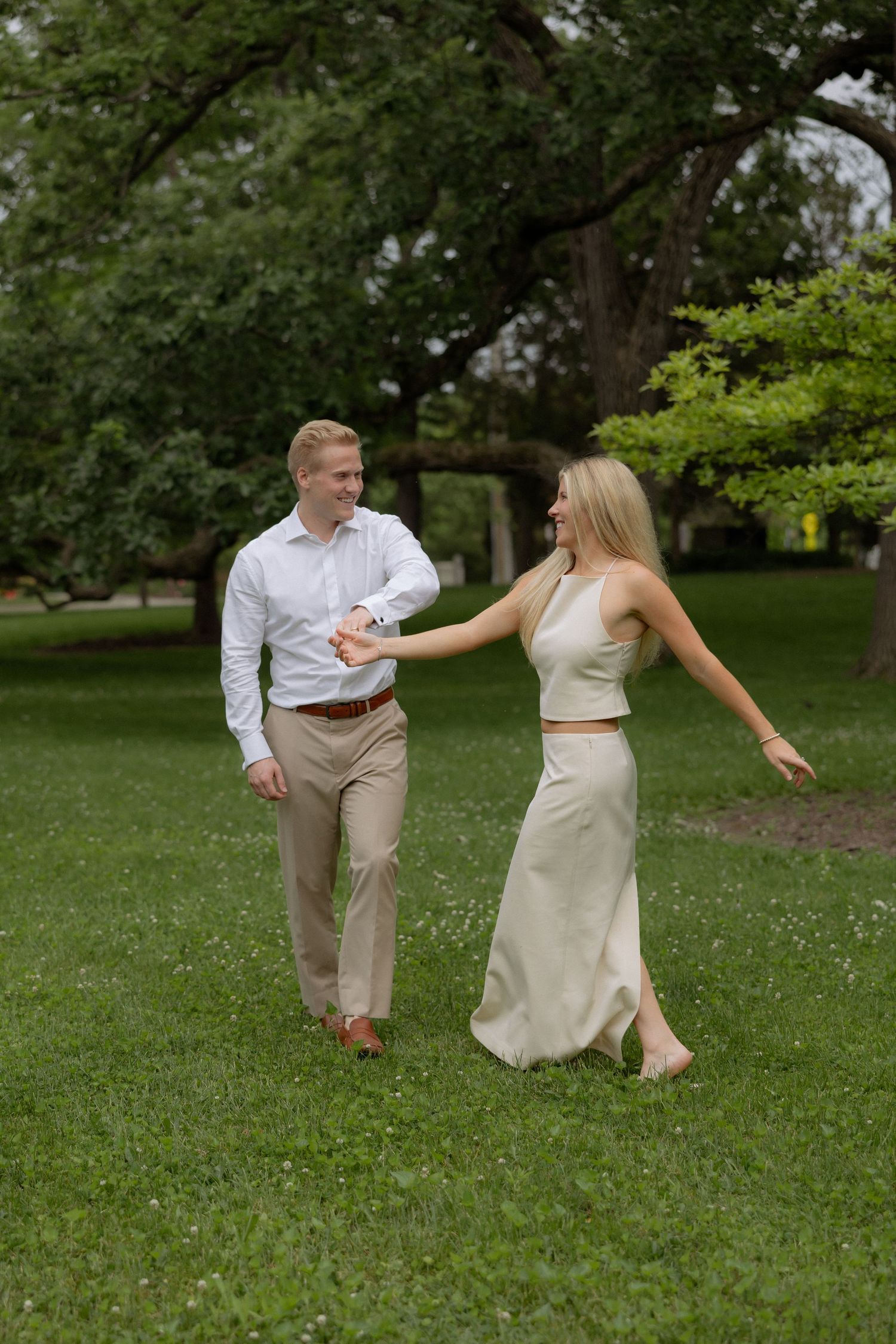 A smiling couple holds hands while walking on lush green grass surrounded by trees in a park.