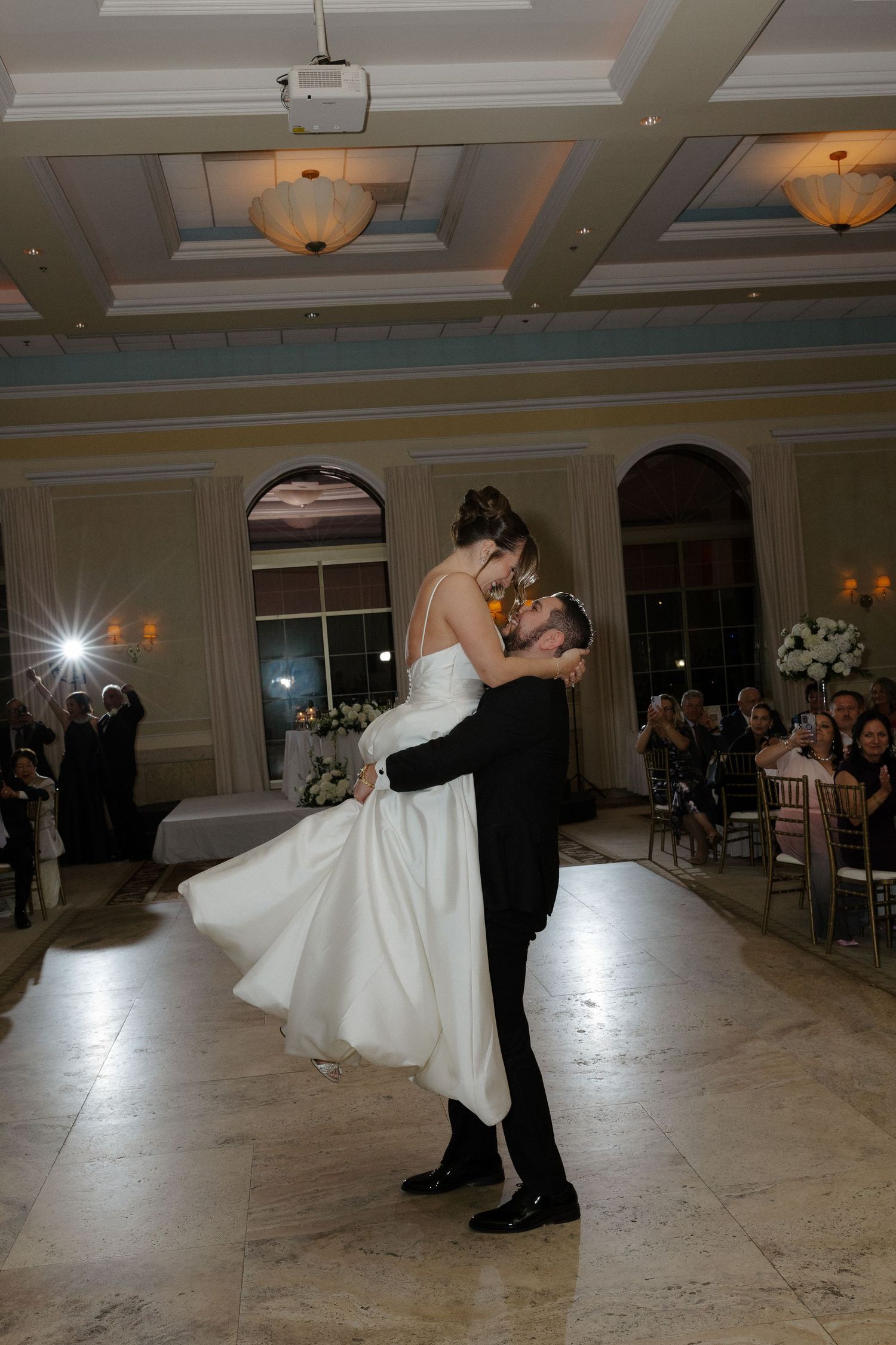 Groom lifts bride in white gown during first dance at elegant wedding reception with guests watching.