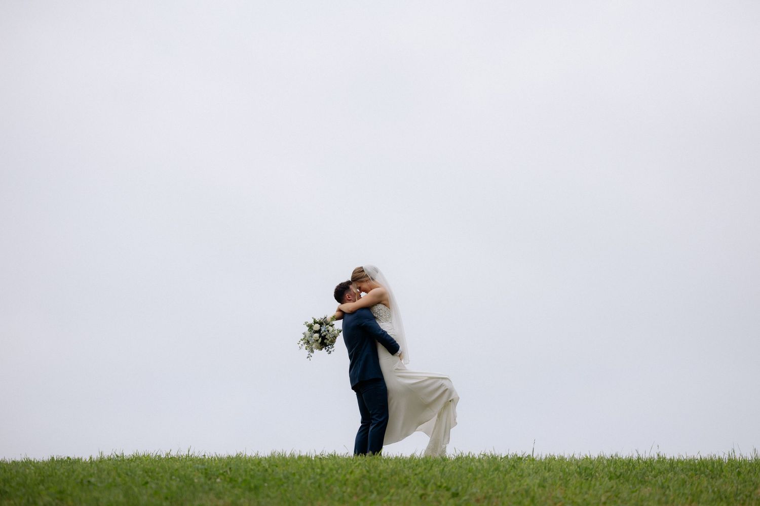 Groom lifting bride in white dress on green hill, romantic wedding couple embrace against gray sky.