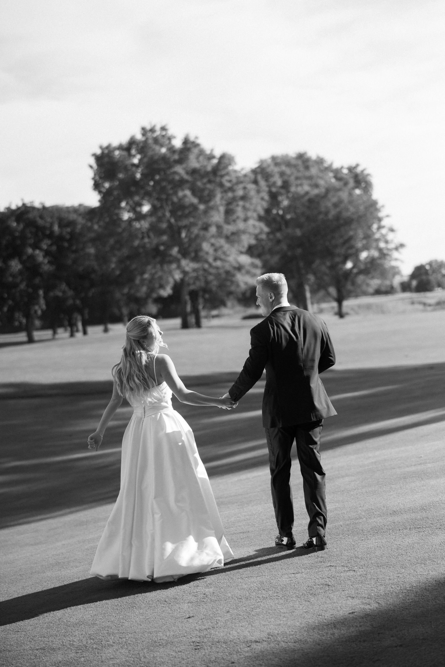 Black and white photo of bride and groom walking hand in hand down a sunlit outdoor path.