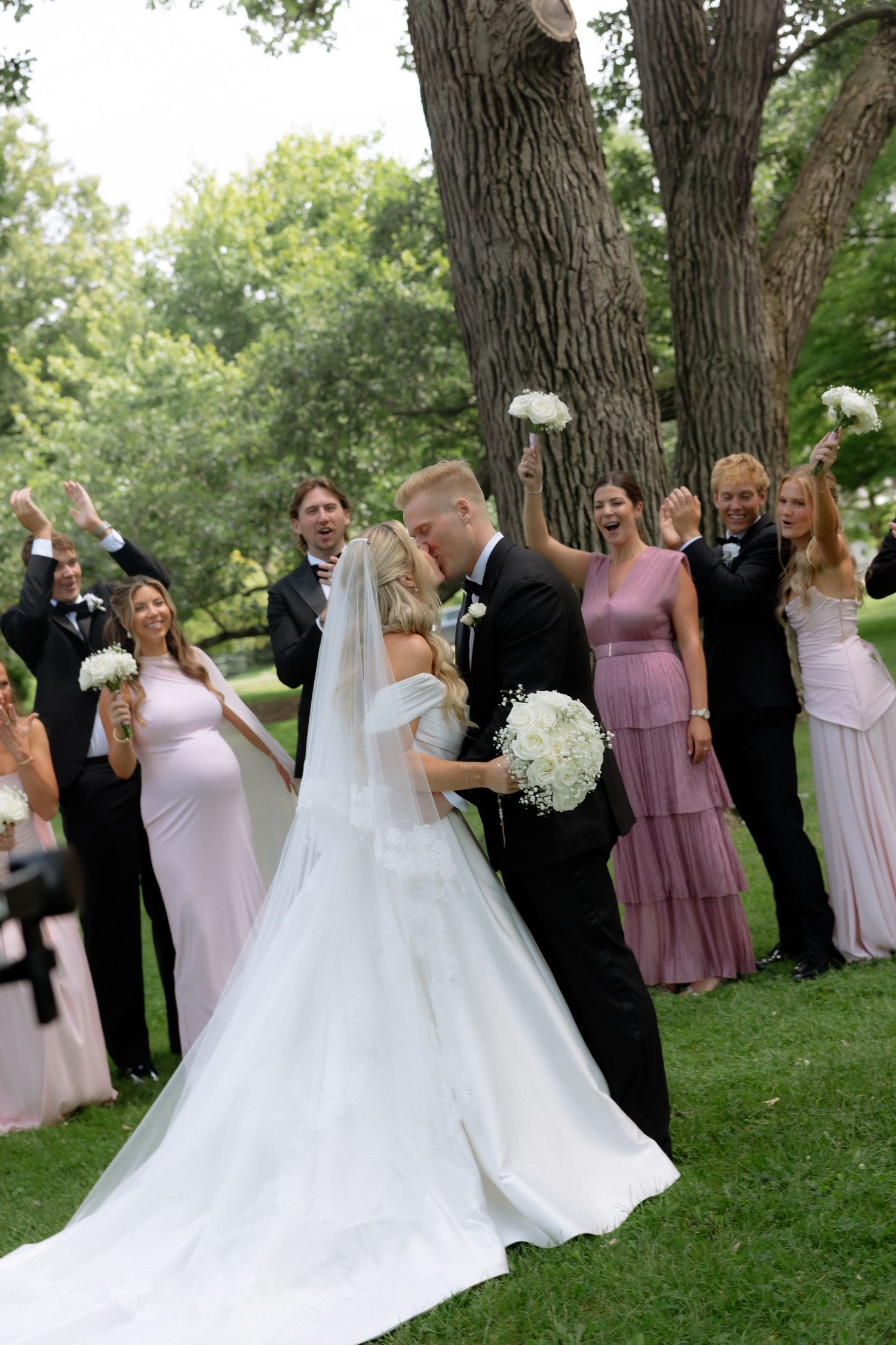Bride and groom share a kiss surrounded by cheering wedding party on lush green lawn.