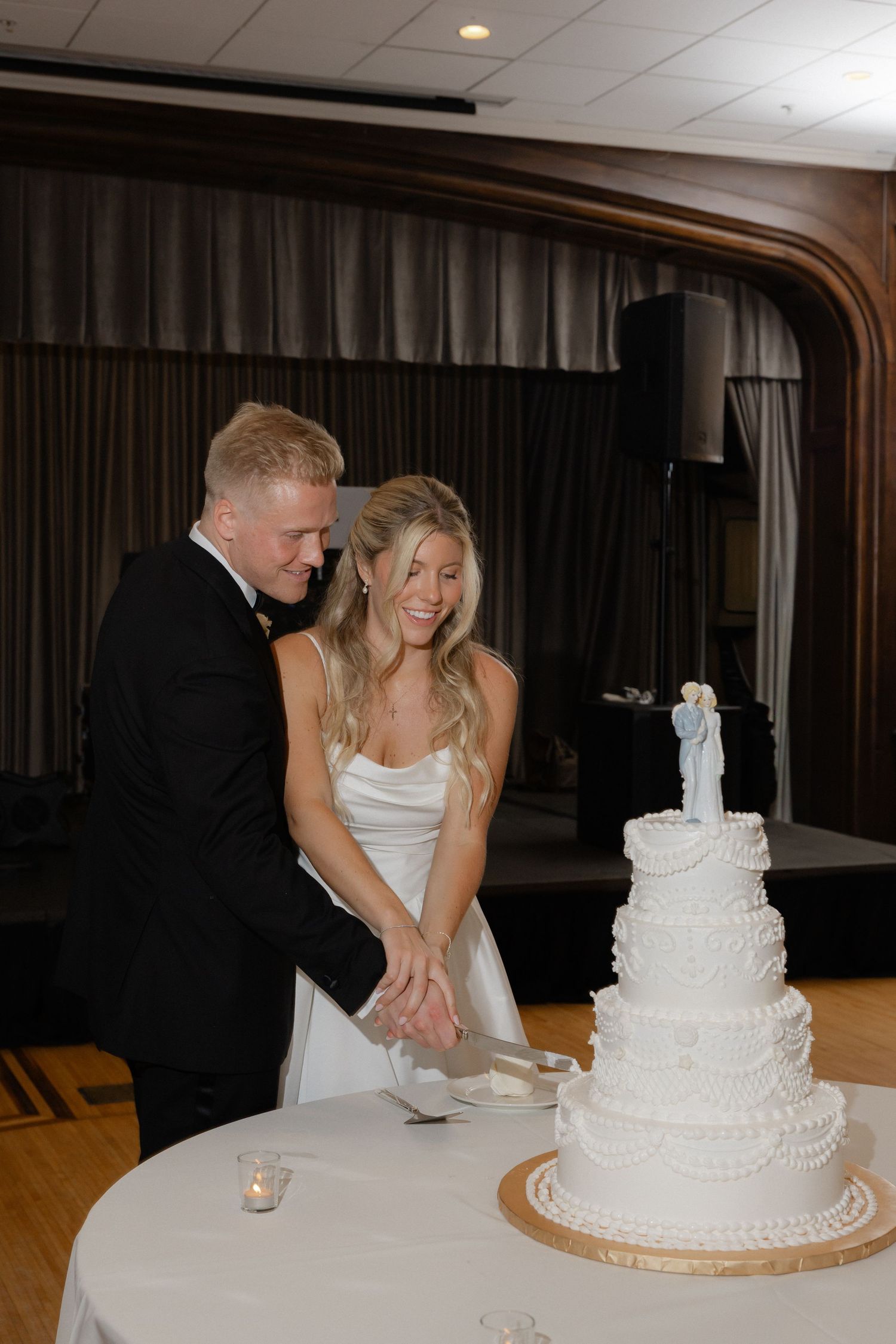 Bride and groom cut elegant four-tier white wedding cake together at indoor reception venue.