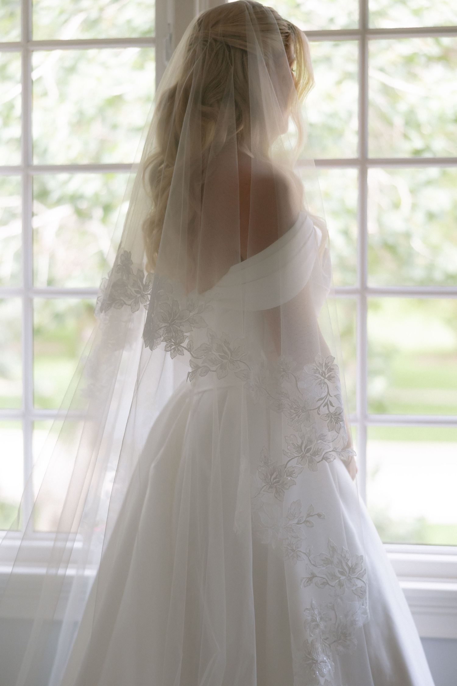 Bride in off-shoulder lace wedding gown and cathedral veil stands before large window in soft light.