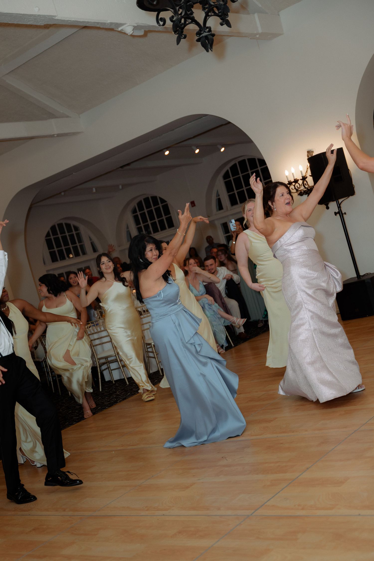 Excited bridesmaids in blue and yellow gowns reaching up to catch the bouquet toss at a wedding reception.