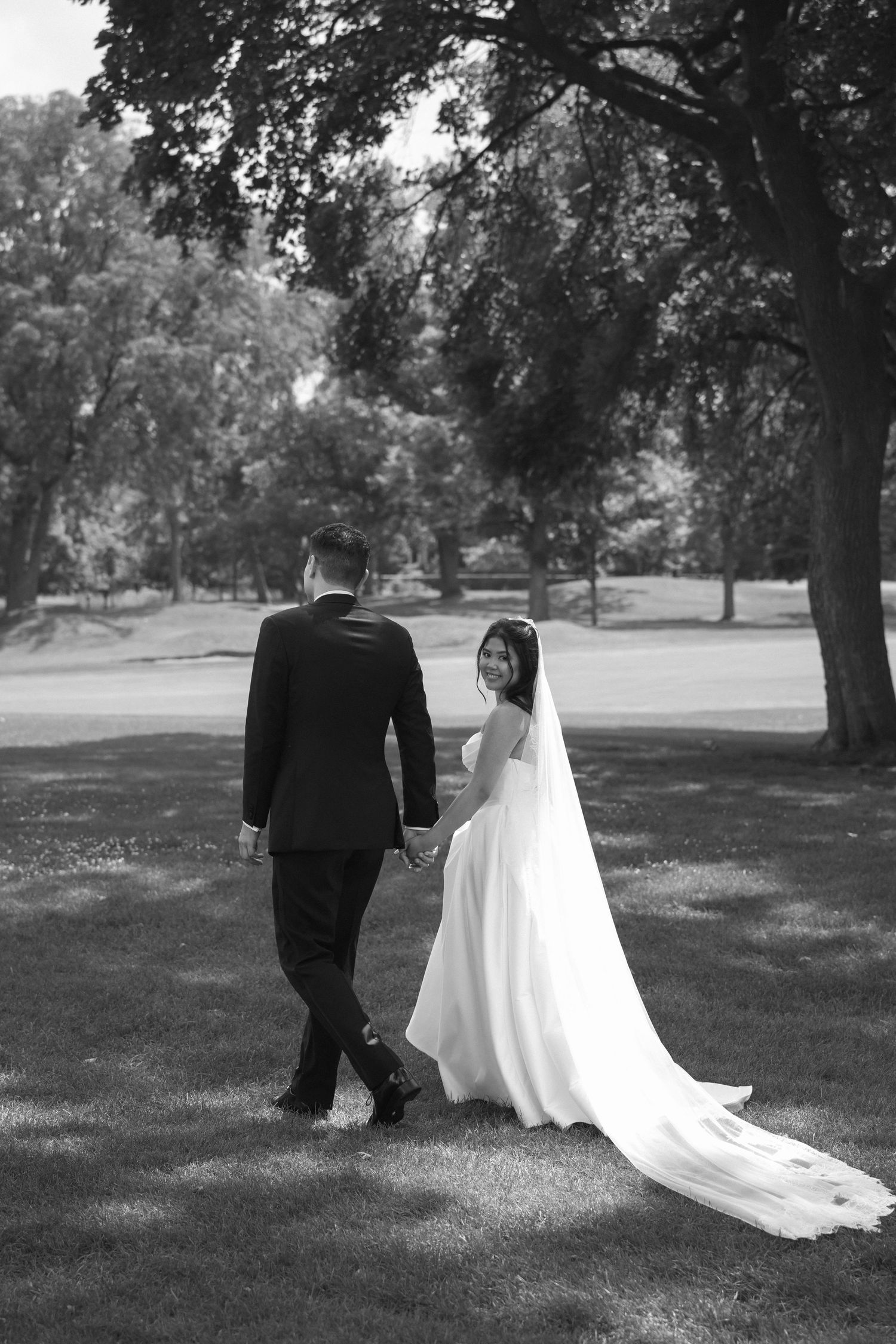 Black and white photo of bride and groom walking hand in hand through a lush green park on their wedding day.