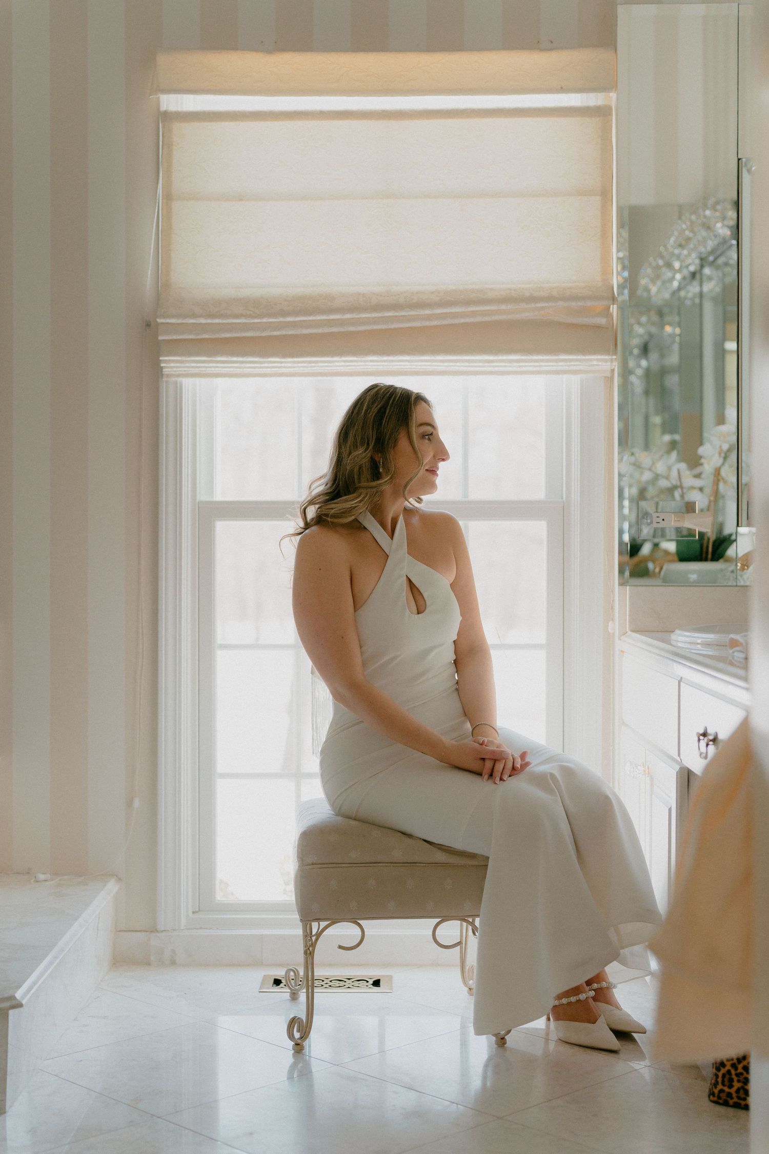 Bride in white halter jumpsuit sitting on bench by window in bright elegant bathroom.