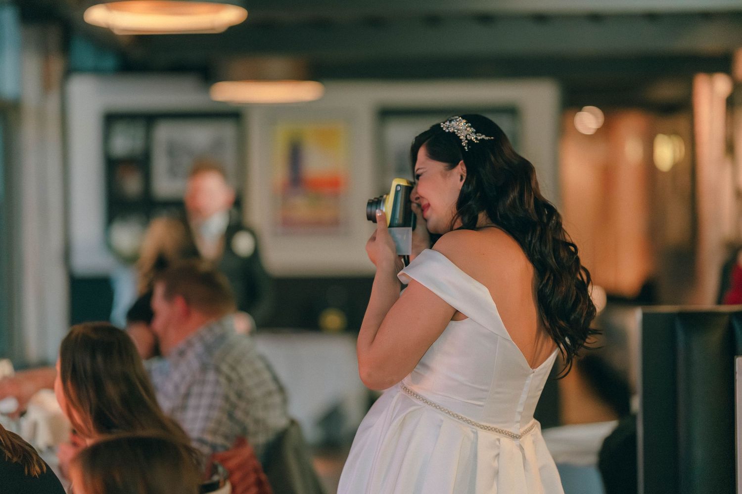 Bride in off-shoulder gown photographs wedding reception guests with vintage camera.