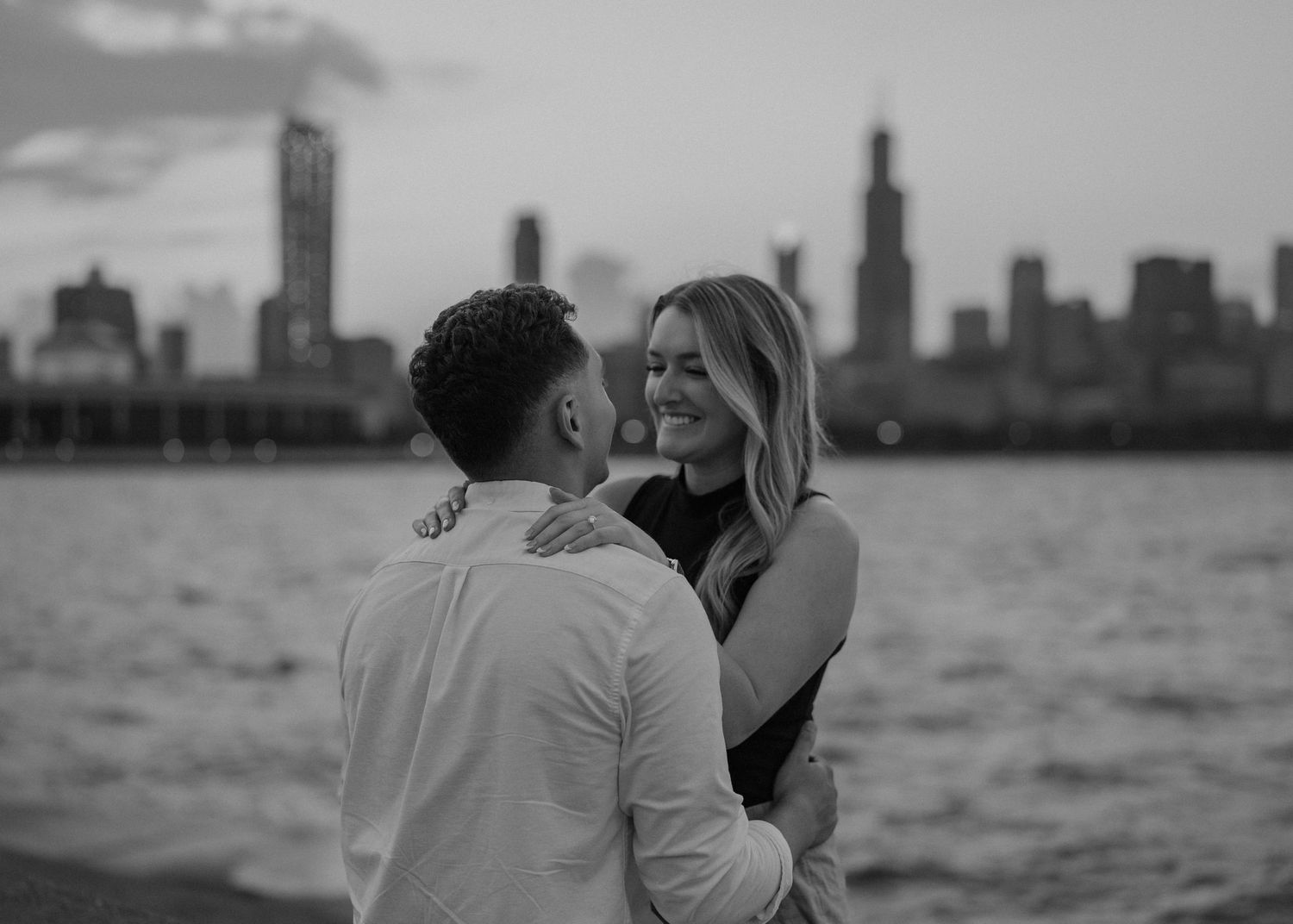 Black and white photo of a couple embracing near Lake Michigan with the Chicago skyline at dusk.