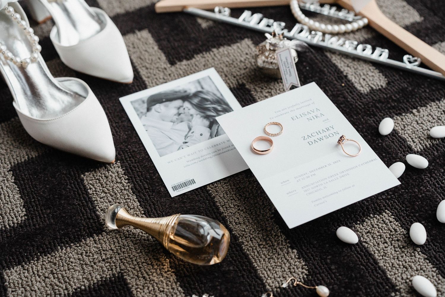 Wedding flat lay with white heels, gold rings, perfume bottles, and invitation on a patterned surface.