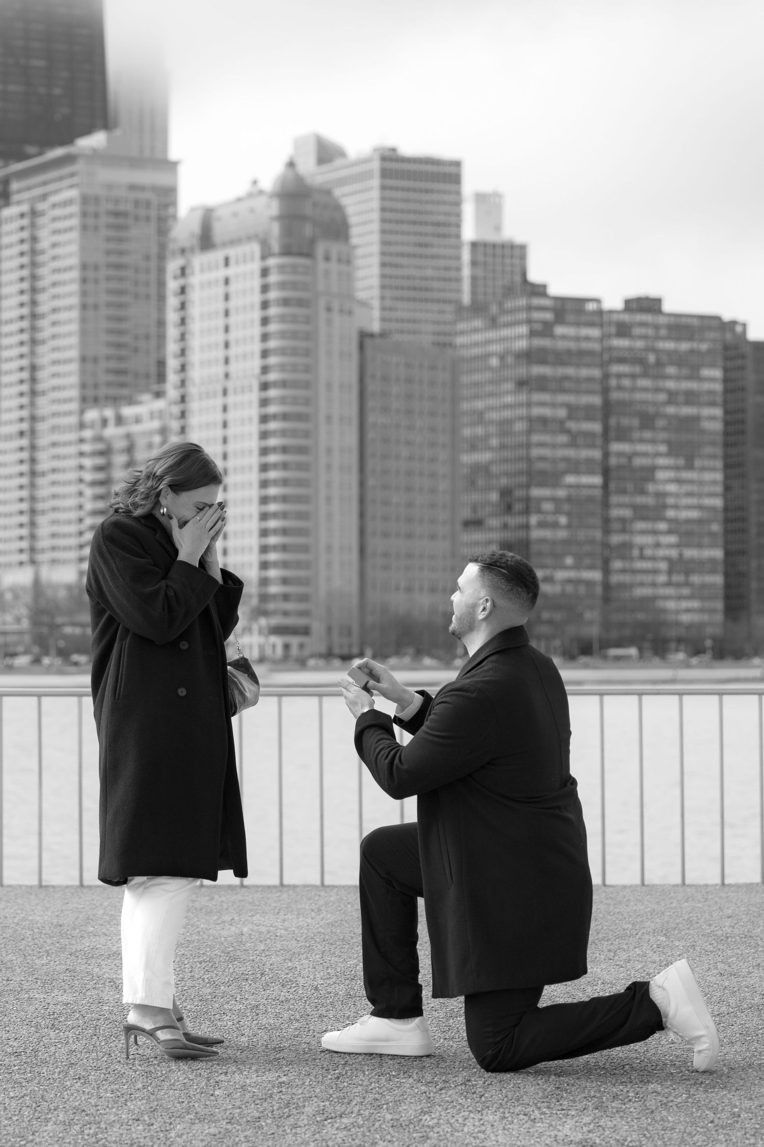 Black and white photo of emotional marriage proposal on Chicago lakefront with man on one knee.